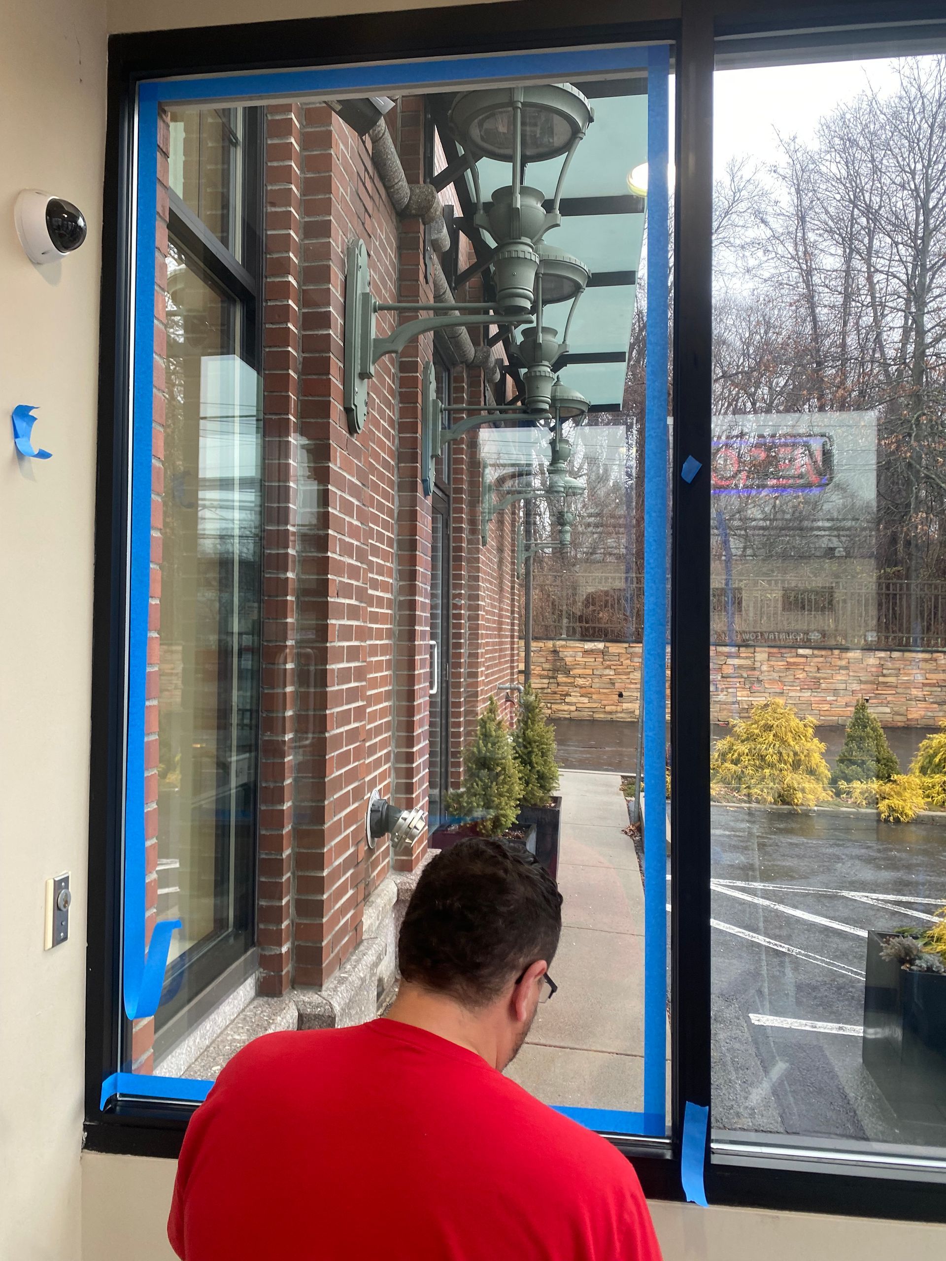 A person in a red shirt applies blue painter's tape to the black frame of a large window inside a brick building.