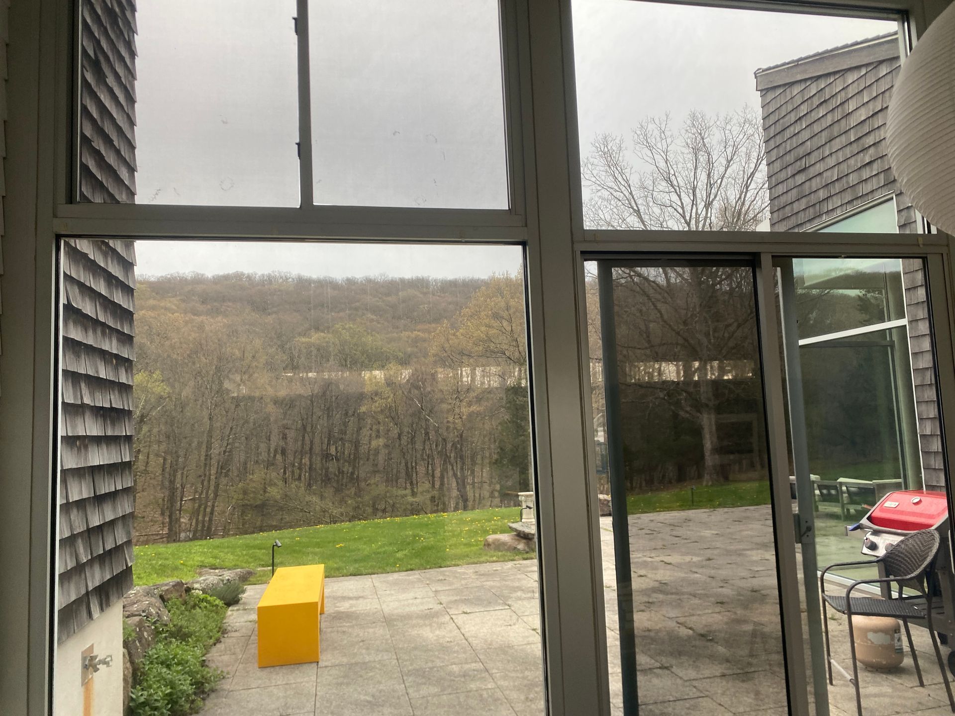View from a window looking out onto a stone patio with a yellow bench, green grass, and trees under an overcast sky.