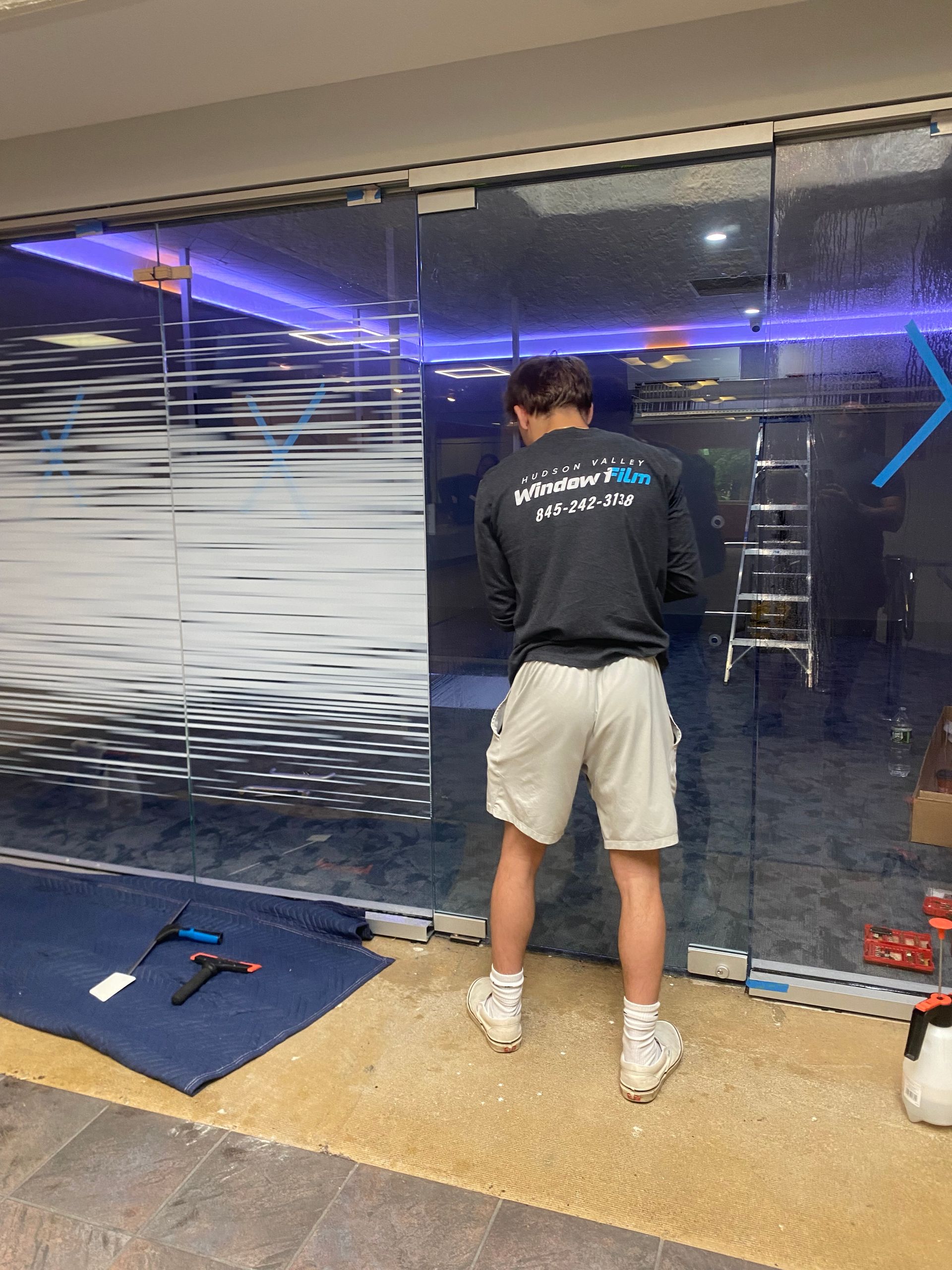 A person in a dark shirt and light shorts stands inside, applying a patterned vinyl film to a large glass storefront.