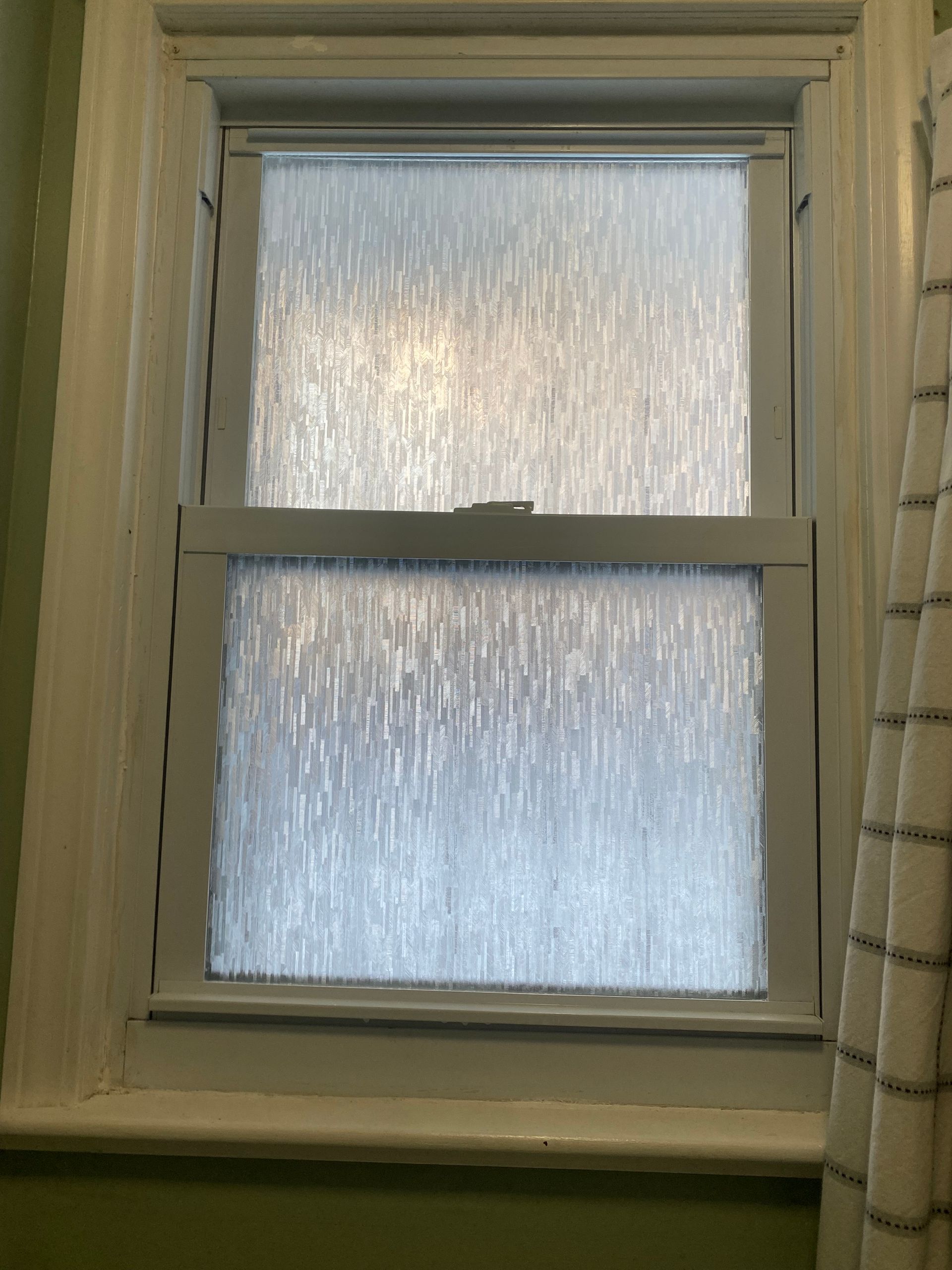 A double-hung window with white frames and privacy film featuring a vertical, textured rain-drop pattern.