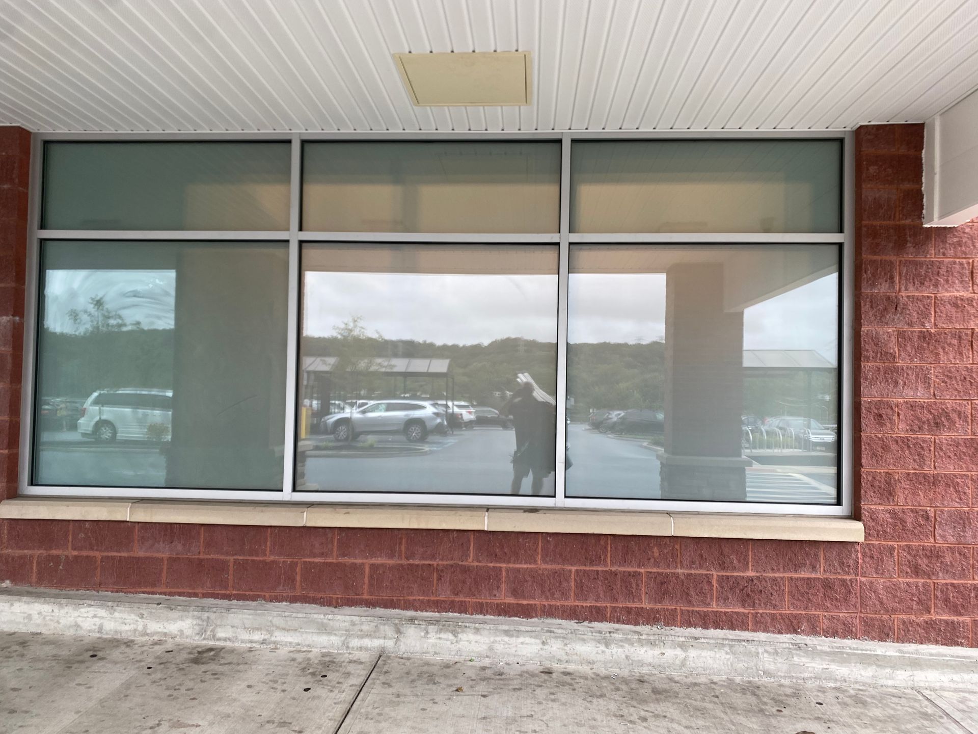 A large window with three panes on a red brick exterior, reflecting a parking lot and a person standing in front.