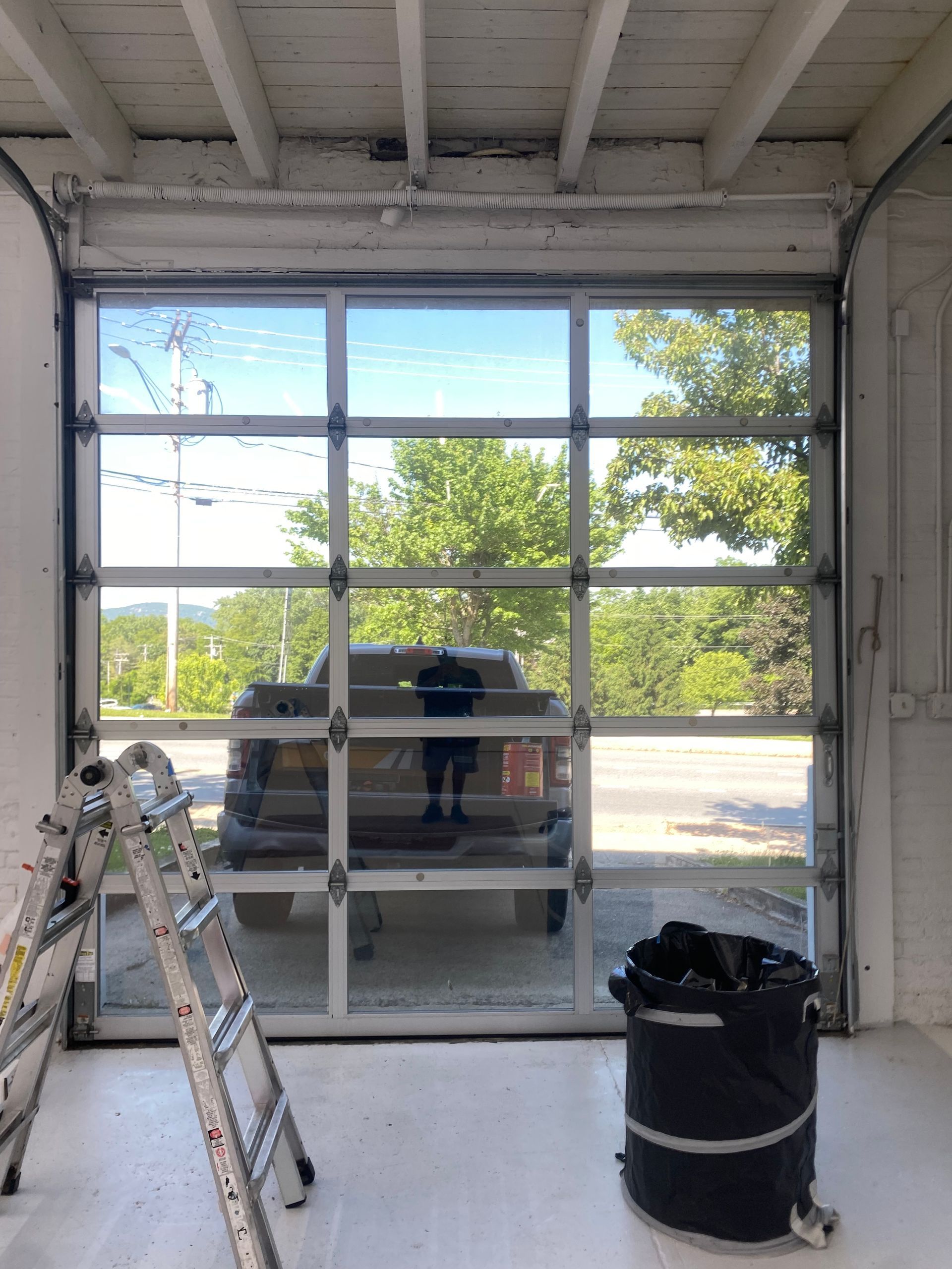 A glass sectional garage door viewed from inside a room, with a vehicle visible outside and a ladder and bin nearby.