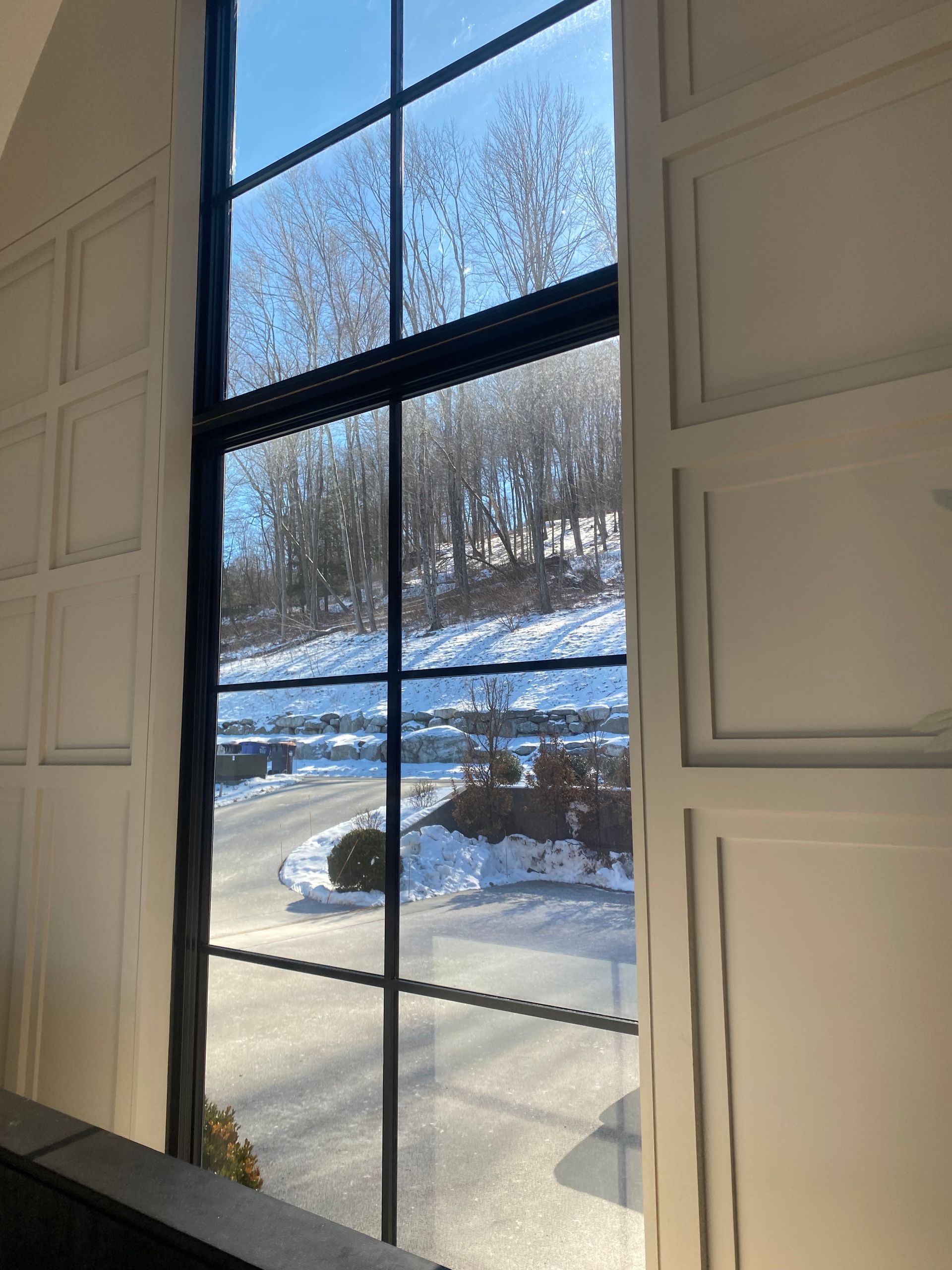 Tall window with black frames looking out onto a snow-covered landscape and trees under a clear blue sky.