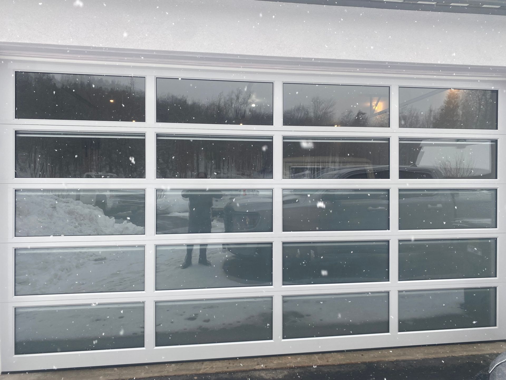 Modern, white-framed glass garage door reflecting a snowy outdoor scene and a vehicle.