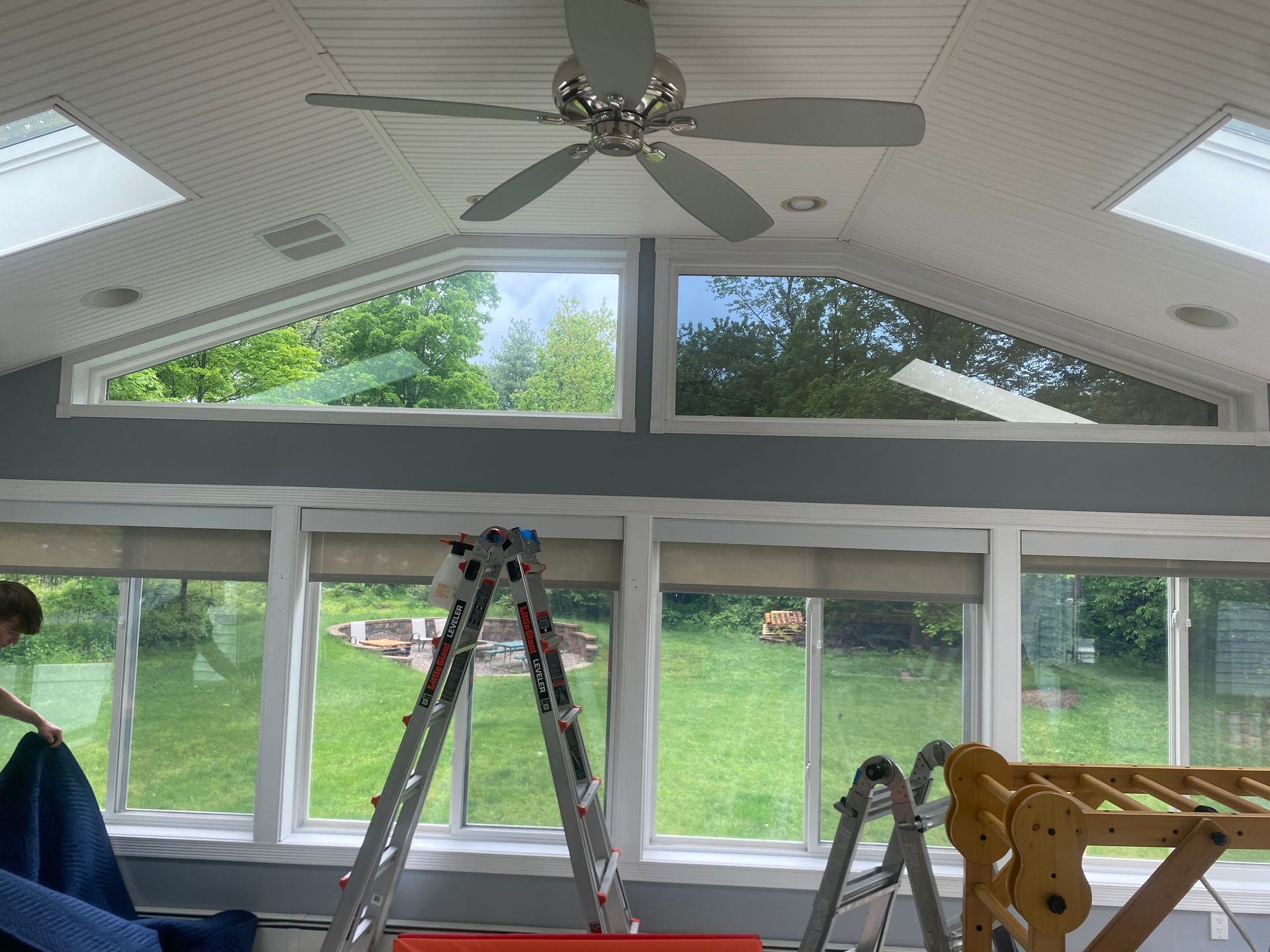 A sunroom interior under construction with gray walls, a ceiling fan, skylights, windows, a ladder, and climbing frame.