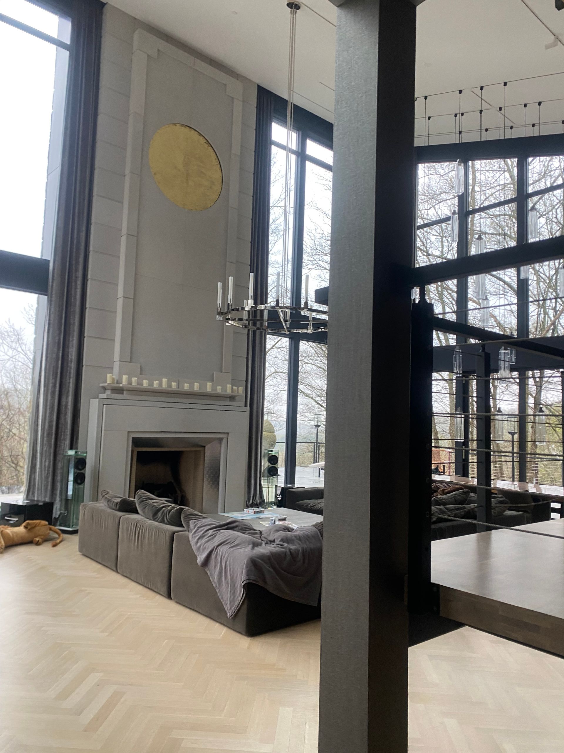 A modern living room with tall windows, a grey fireplace featuring a gold circular accent, and a large sofa.