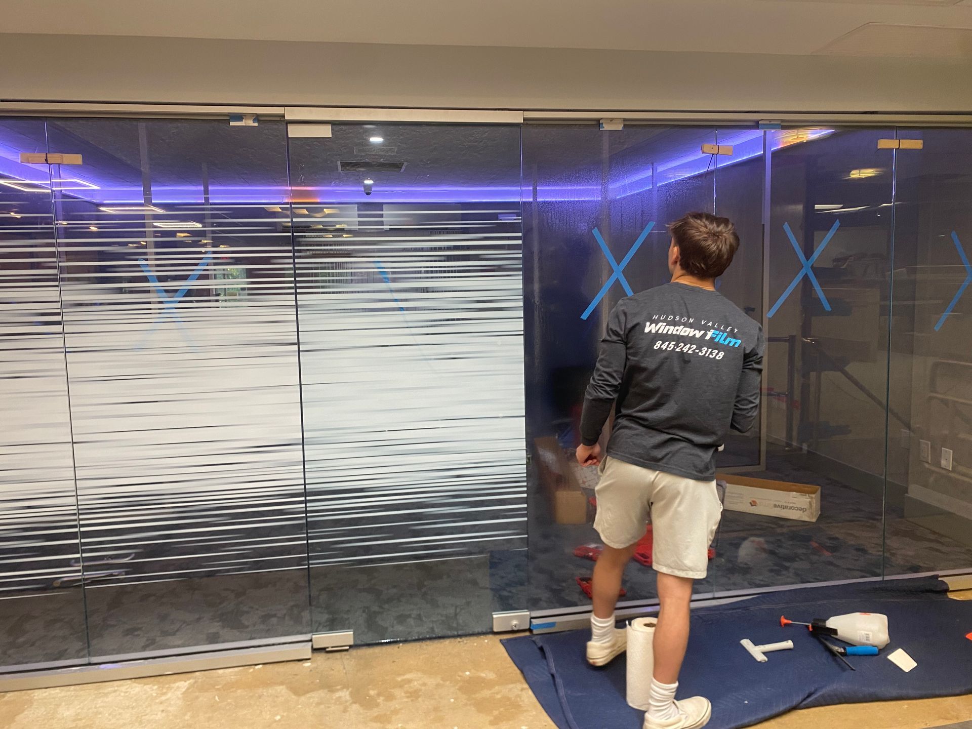 A worker applies a frosted, wavy-patterned film to a large glass office partition, with tools laid on a blue drop cloth.