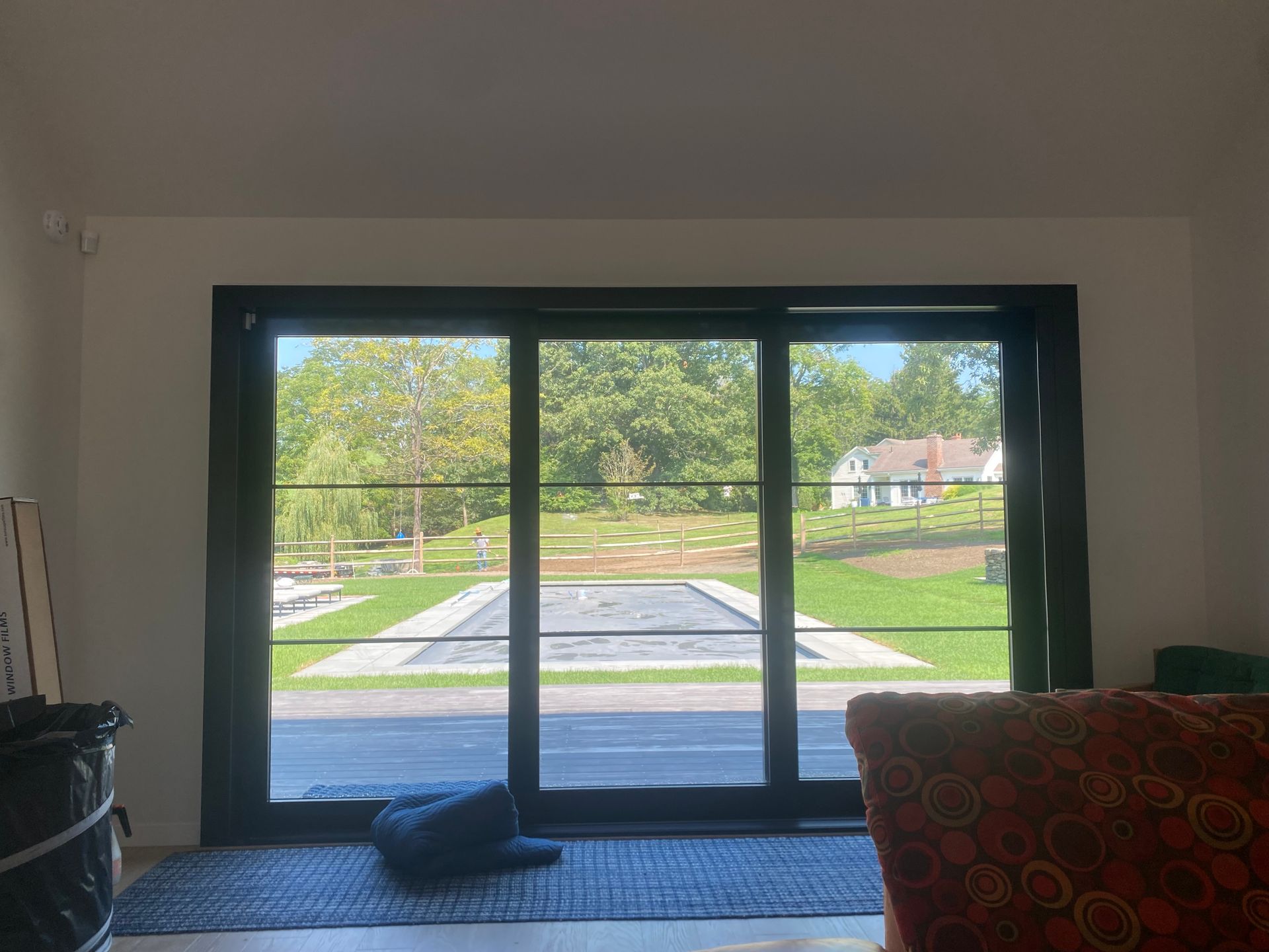 A large black-framed sliding door overlooks a sunny backyard with a pool, grassy hills, and a house in the distance.