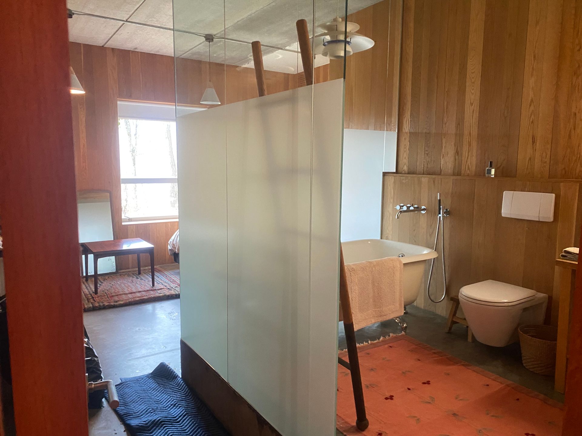 A bathroom and bedroom space divided by a frosted glass partition, featuring a clawfoot tub, a toilet, and wood paneling.