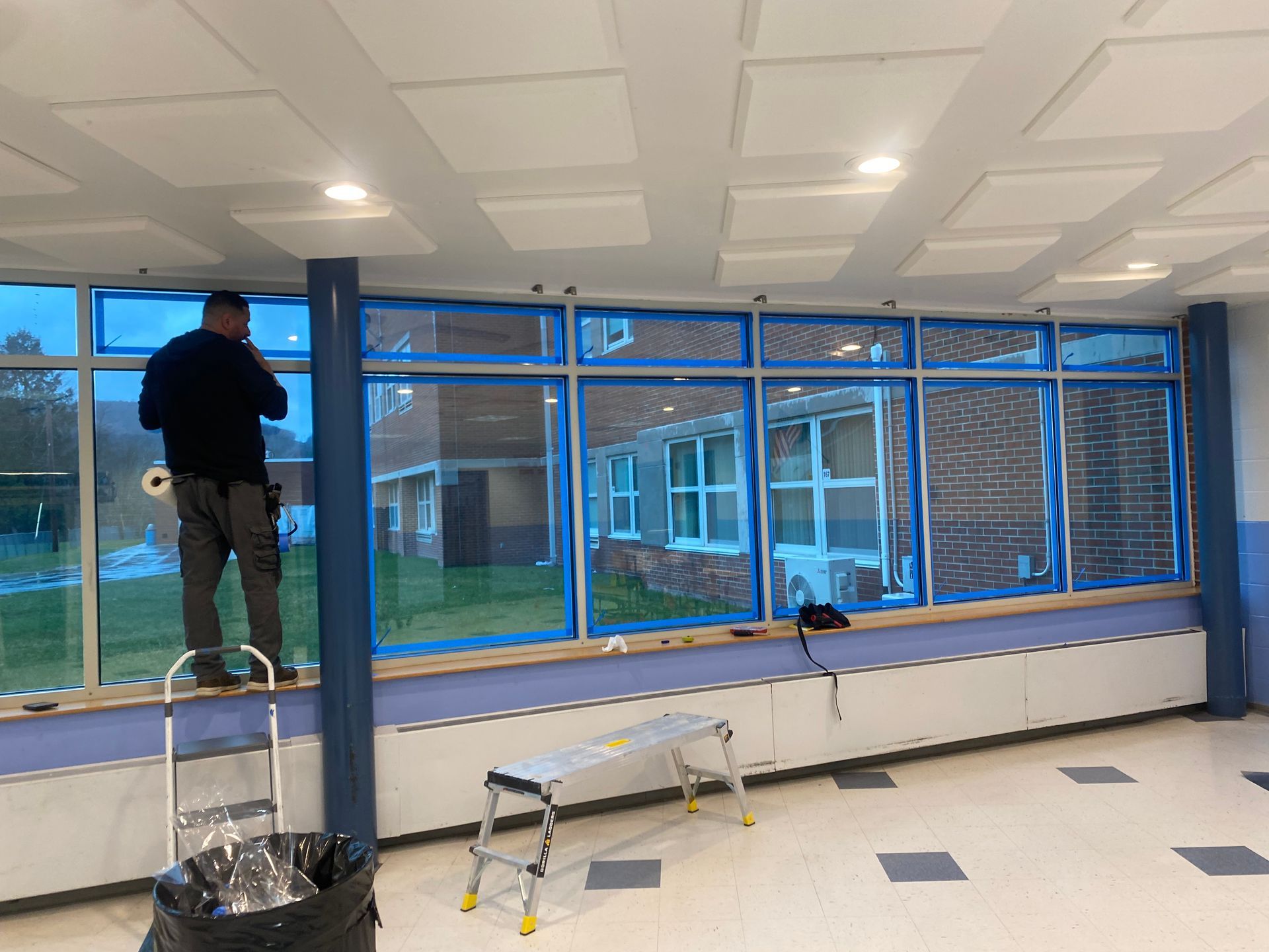A person stands on a ledge inside a room, applying blue masking tape to the frames of a row of windows.