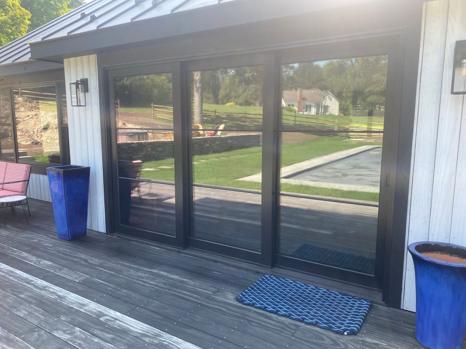 Black sliding glass doors on a wooden deck, flanked by two blue planters, with a grassy lawn and swimming pool reflected.