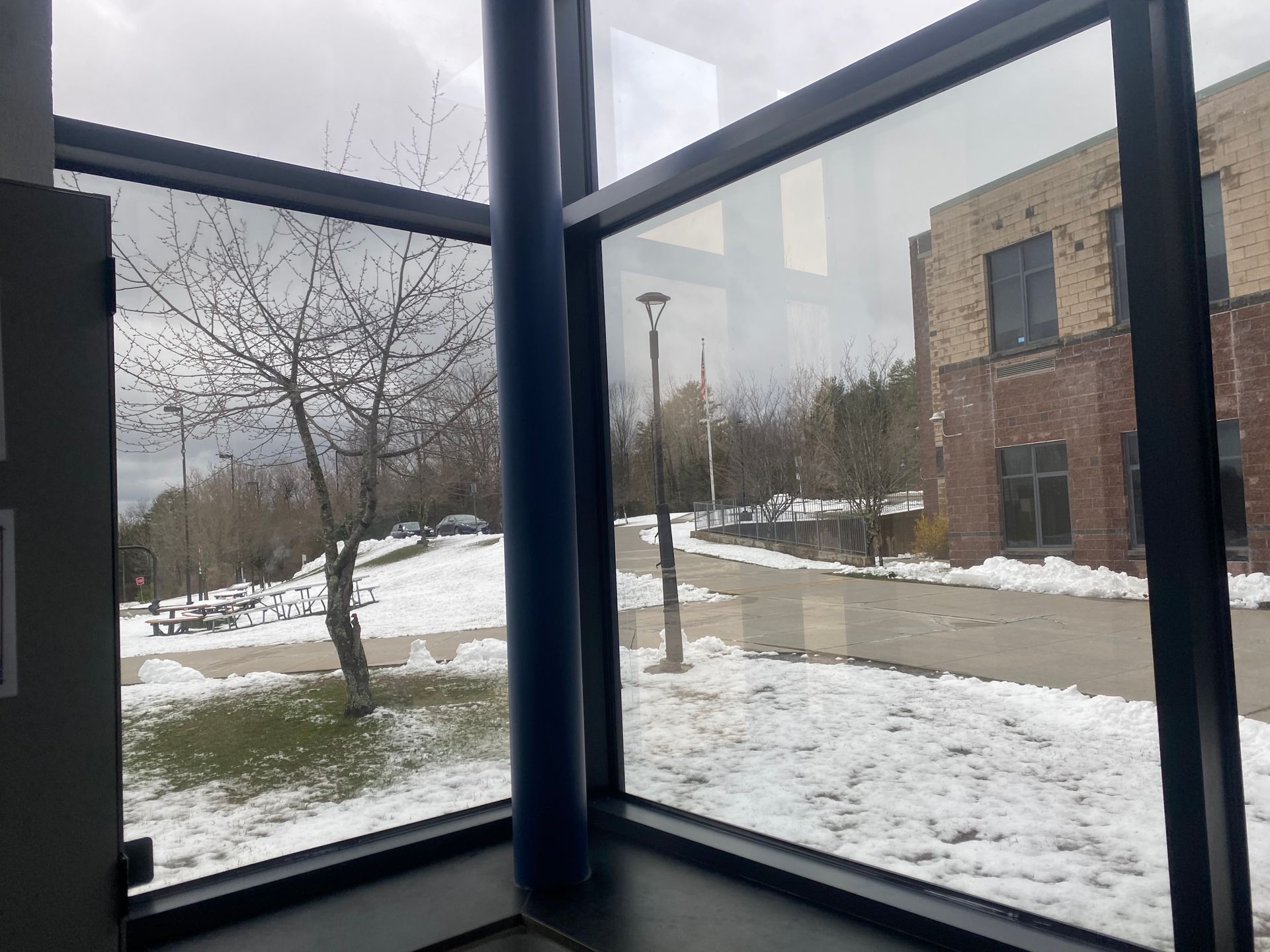 A view through a large window showing a snow-covered outdoor area with a tree and a brick building under a cloudy sky.