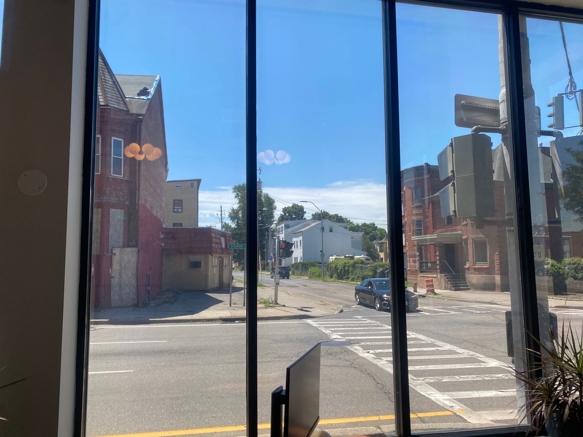 A view through a window showing a street intersection with brick buildings, a crosswalk, and a car on a sunny day.