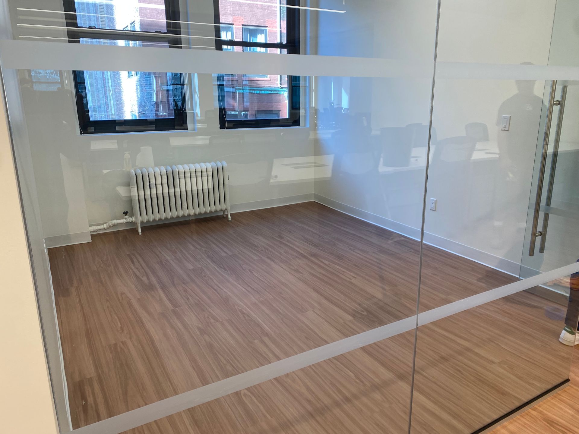 A small, empty office space with wood-look flooring and a white radiator, viewed through a glass wall.