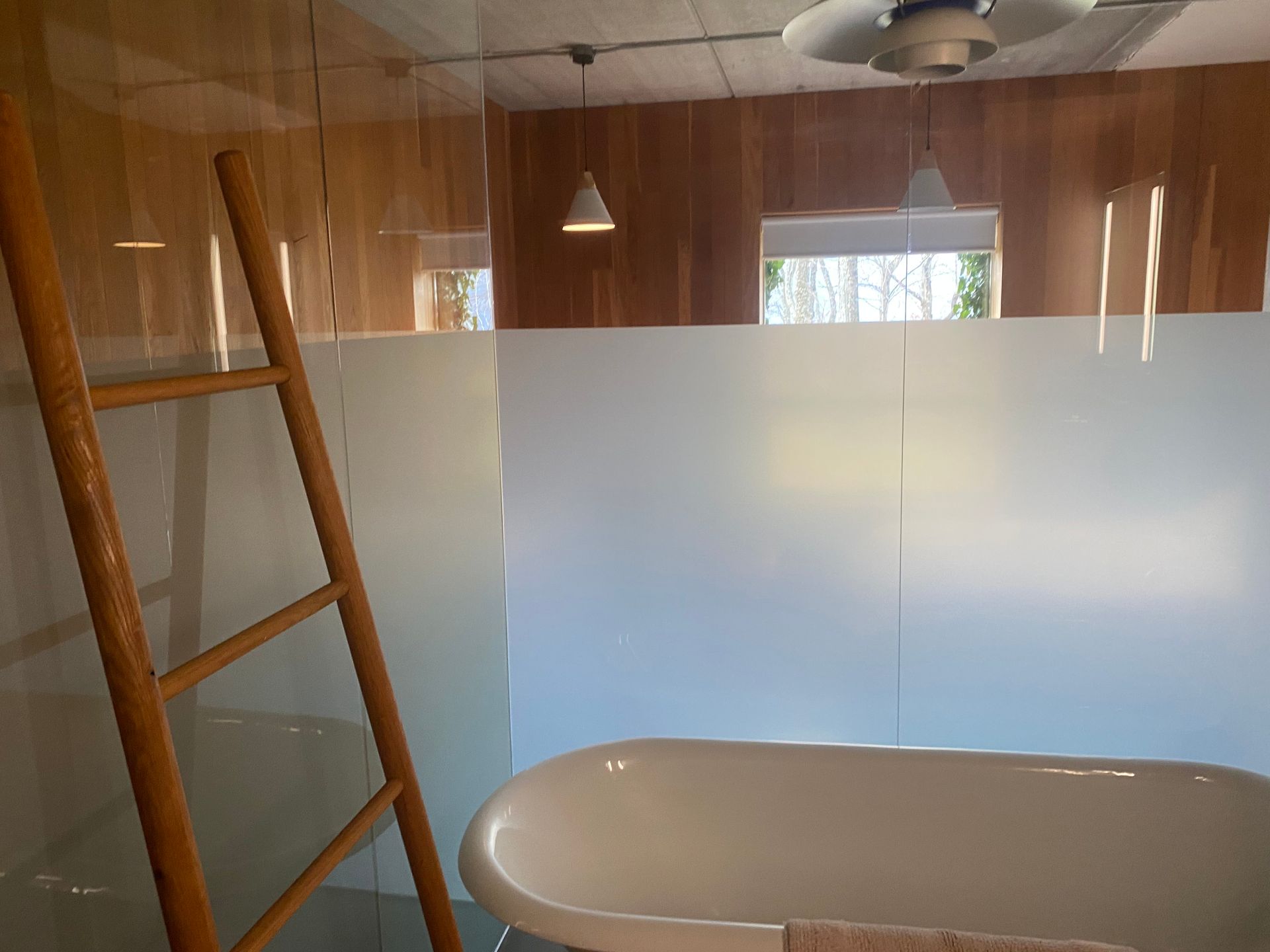 A white clawfoot bathtub sits in a modern bathroom with a wood-paneled wall, hanging pendant lights, and a wooden ladder.