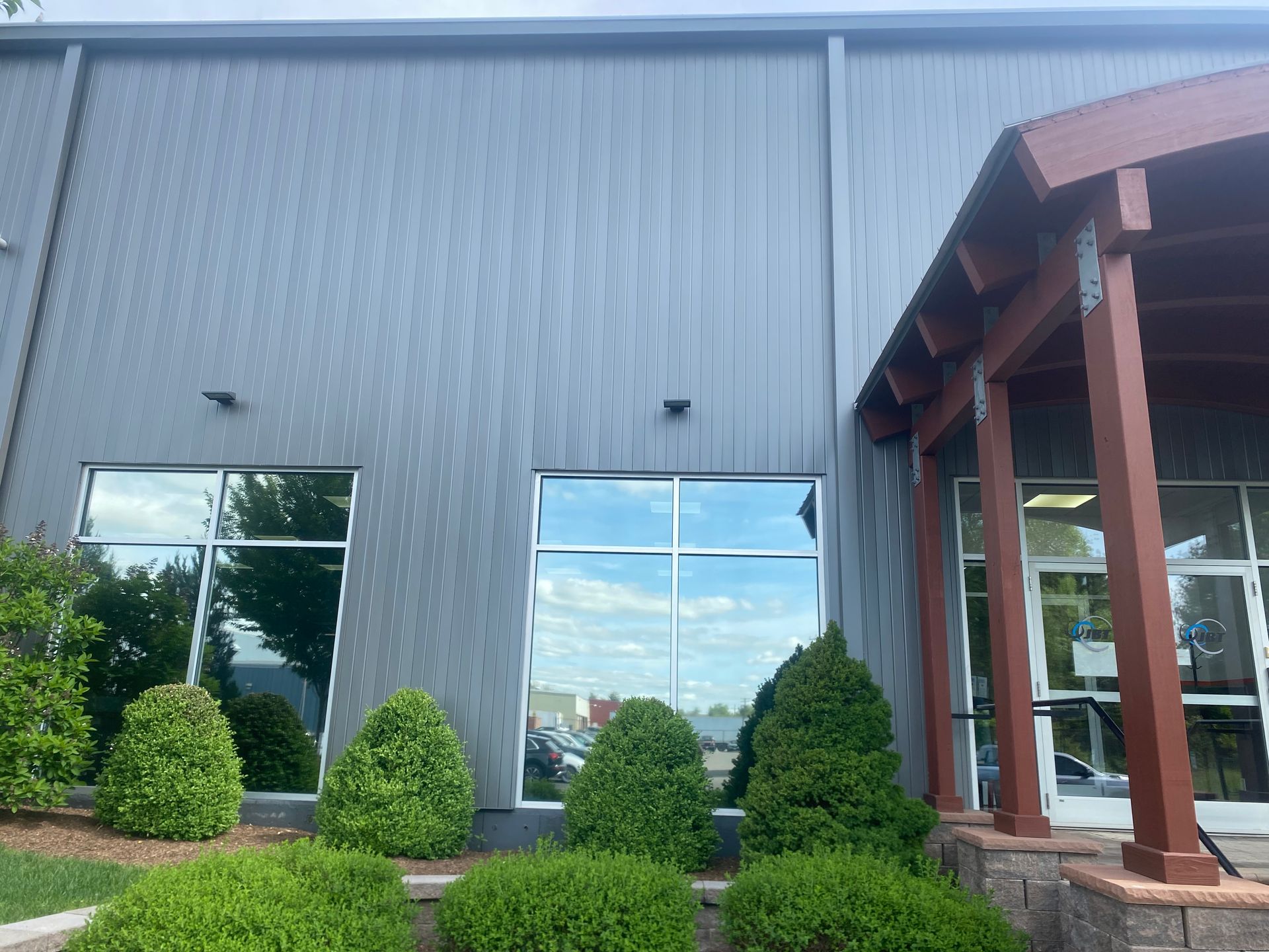 A modern building with gray metal siding, two reflective windows, and a covered wooden entryway with small green shrubs.