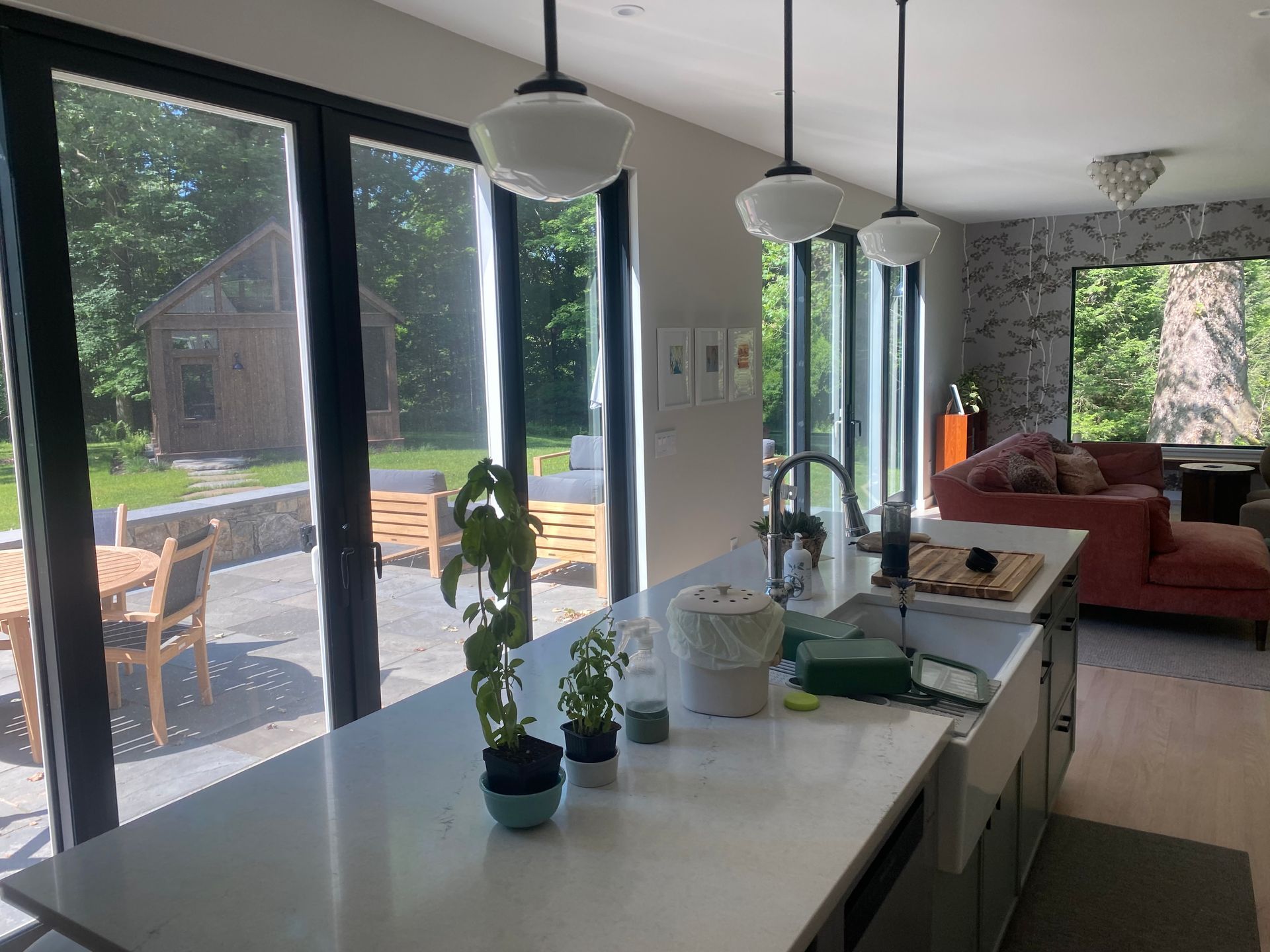 A bright modern kitchen with a white island, hanging pendant lights, and large glass doors looking onto a patio and woods.
