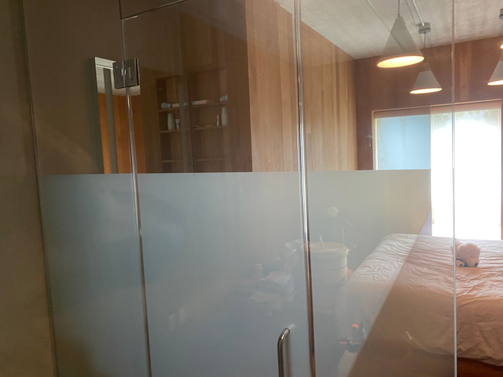 A frosted glass shower partition in a room with wood paneling and a bed visible through the transparent upper portion.