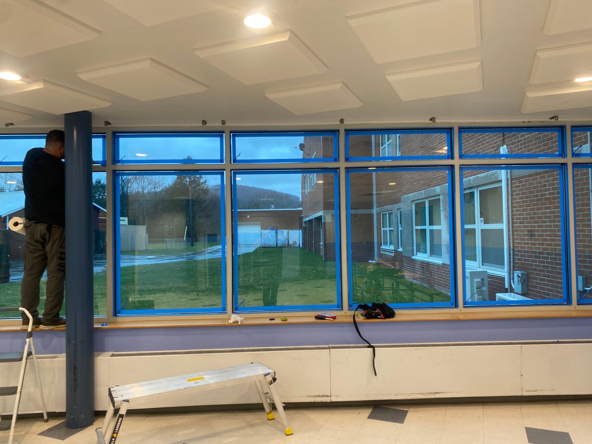 A worker installing blue-tinted privacy film on a large row of windows in an office or classroom.