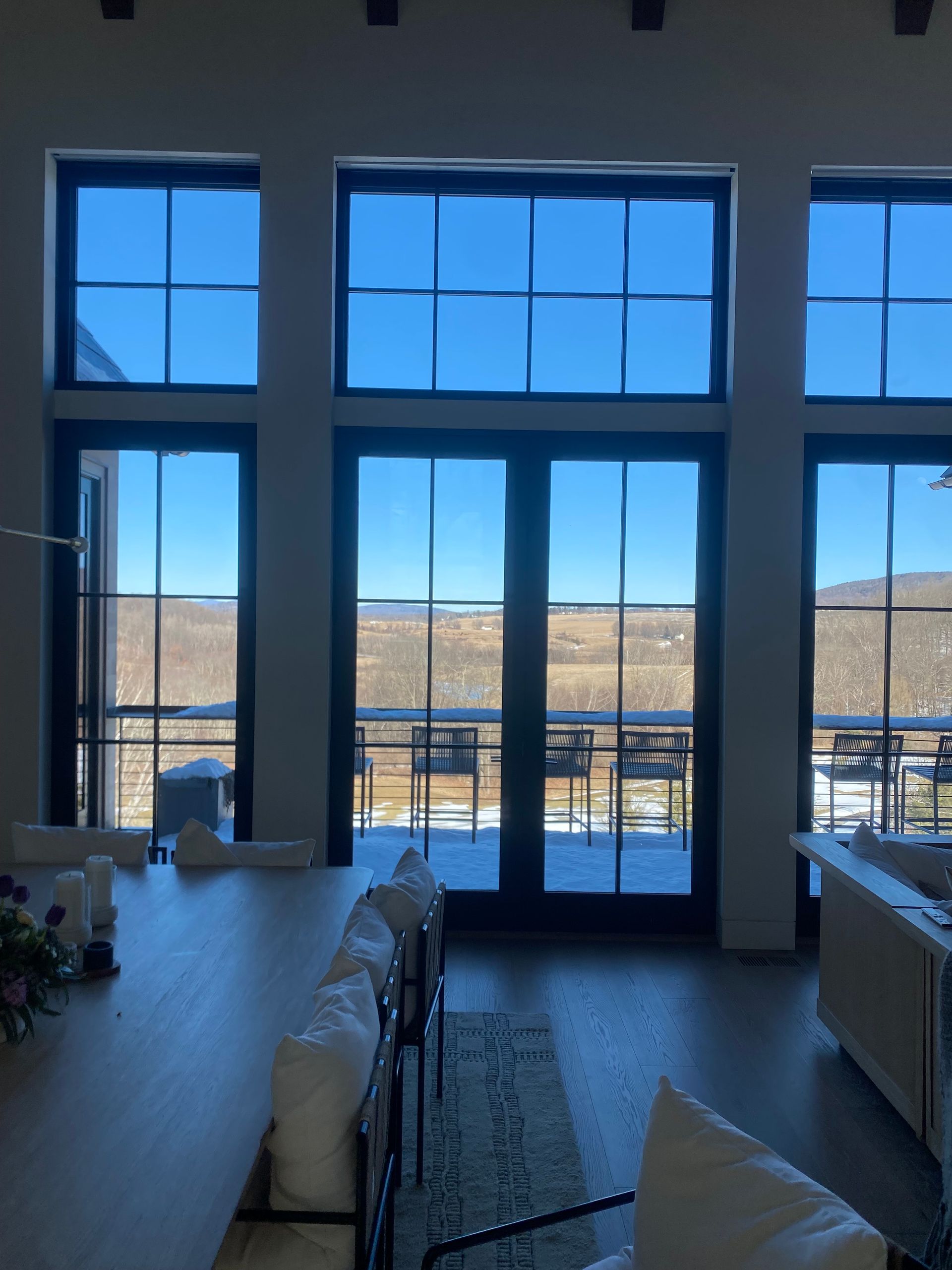 Large floor-to-ceiling windows look out onto a snowy, sunlit landscape from a bright dining area with white chairs.