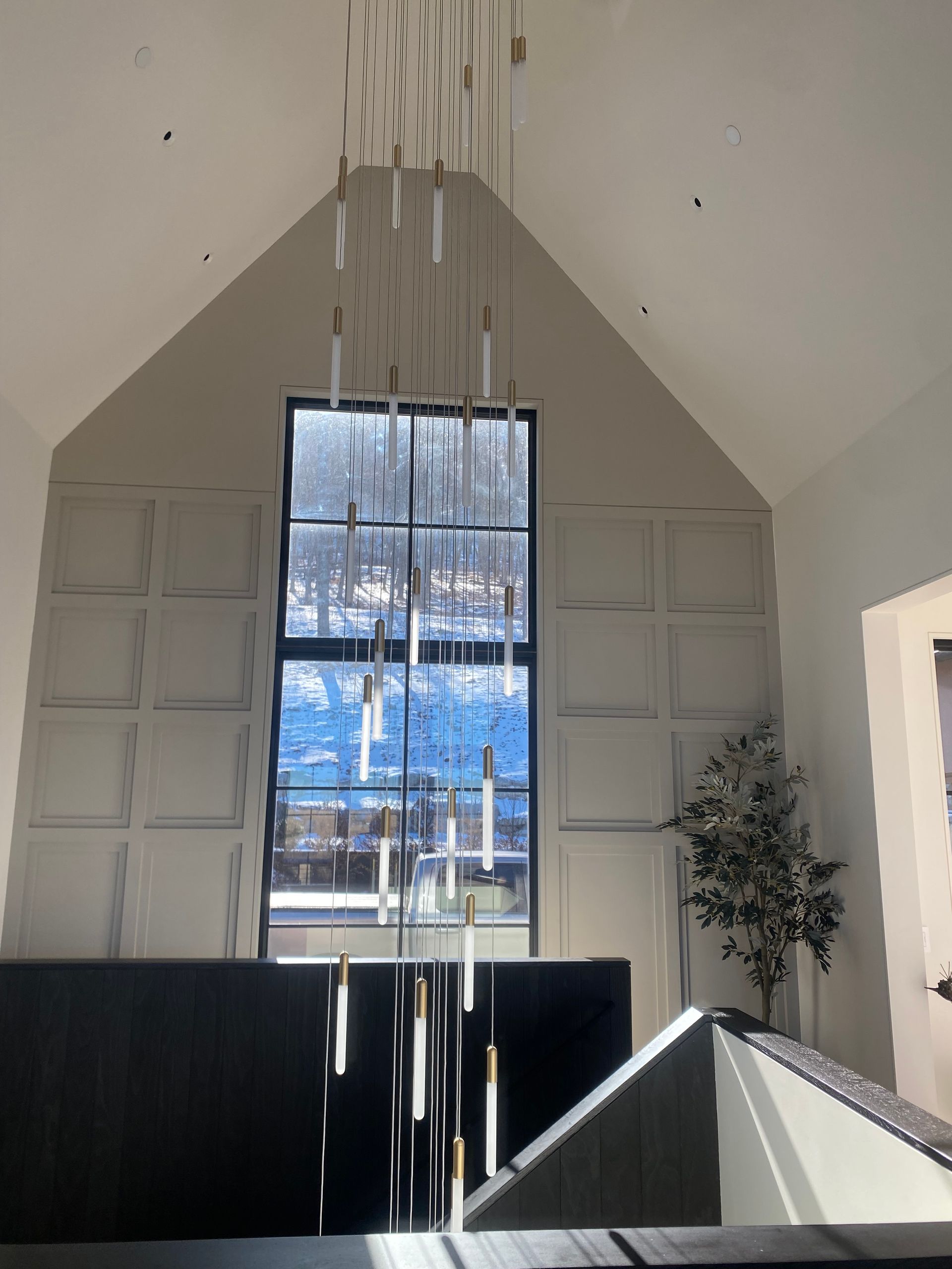 A tall, multi-pendant chandelier hangs before a large window with paneled walls in a high-ceilinged, sunlit room.
