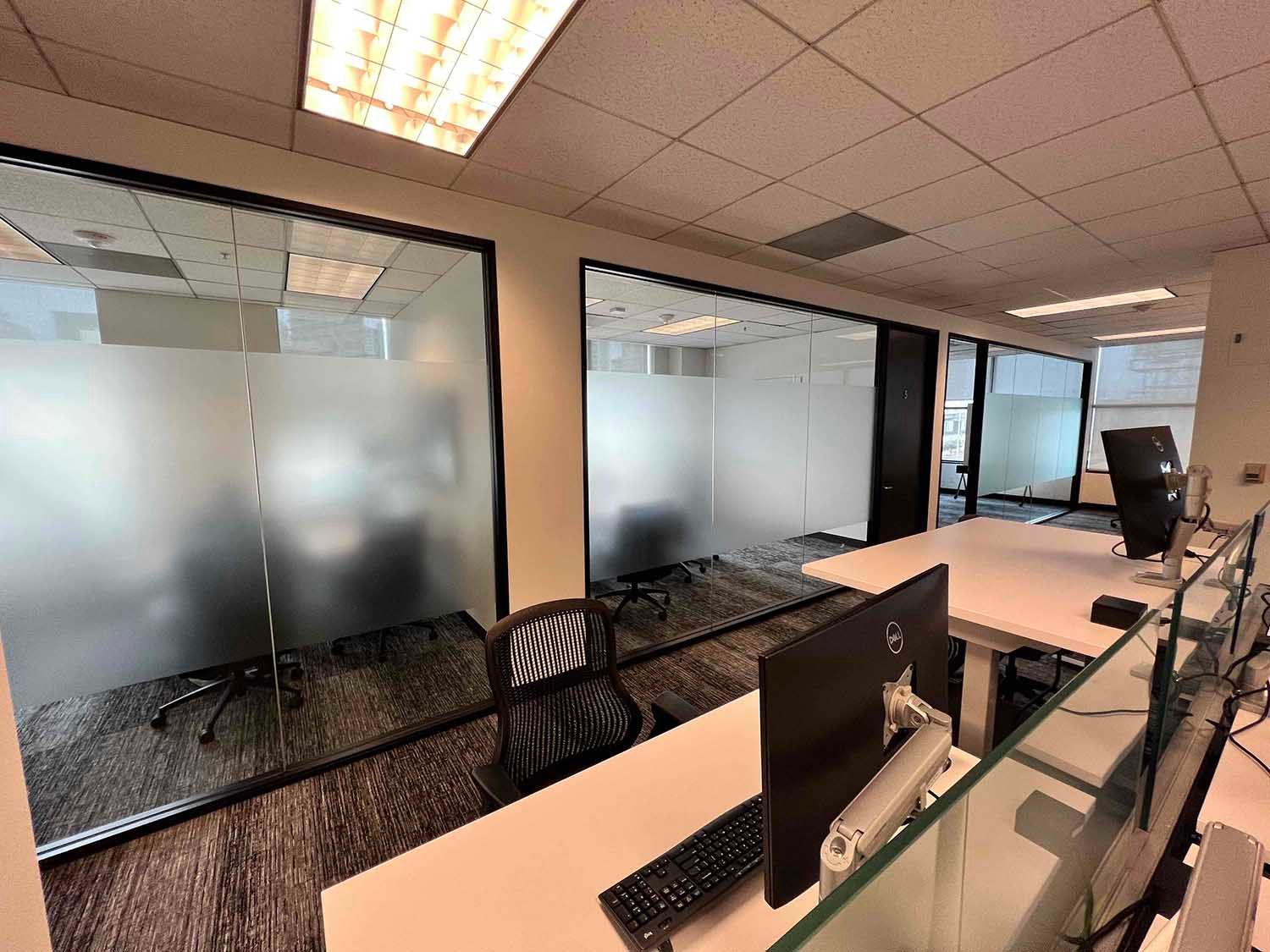 A modern office interior featuring several enclosed workstations with frosted glass walls and white desks.