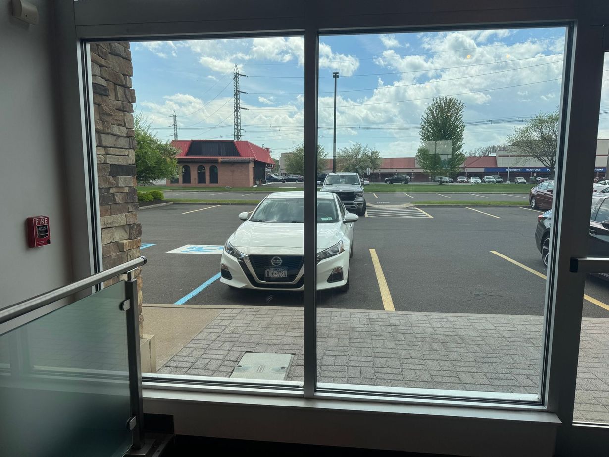 A view through a glass door looking out onto a parking lot with a white sedan parked in a handicapped spot.
