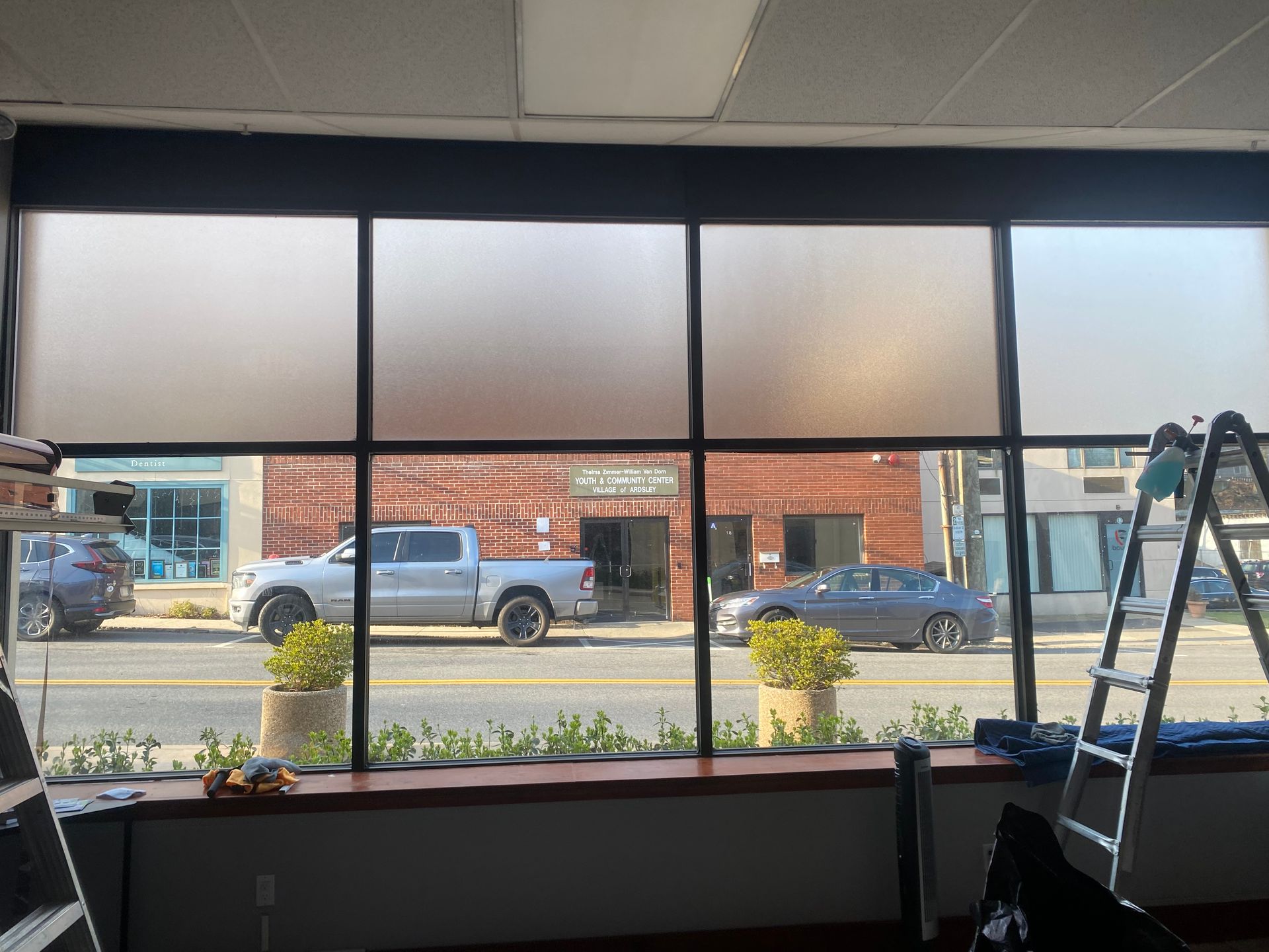 A street view through four large, frosted office windows, with construction ladders visible on both sides of the frame.