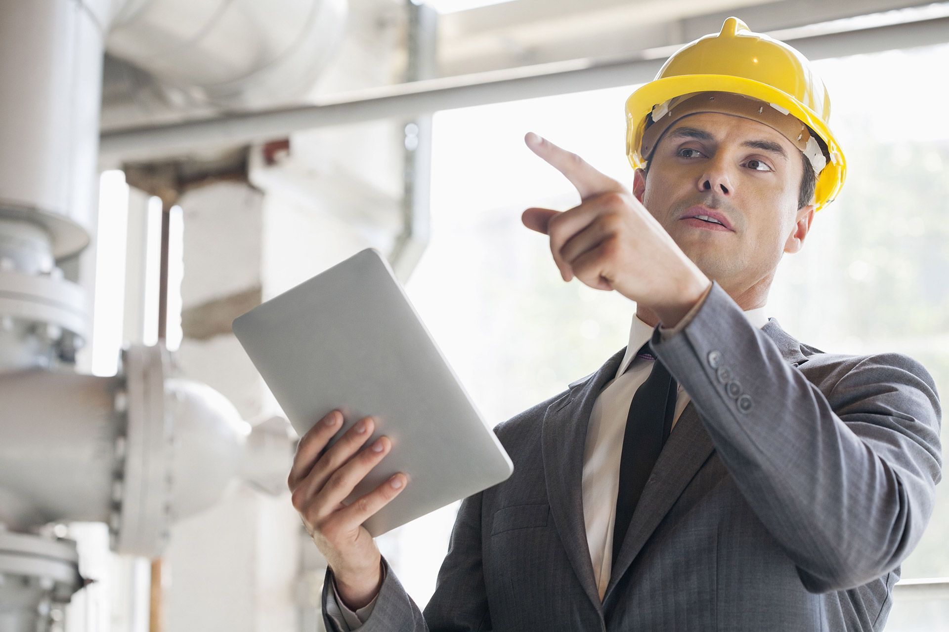A man in a suit and hard hat is holding a tablet and pointing at a part of an industrial facility