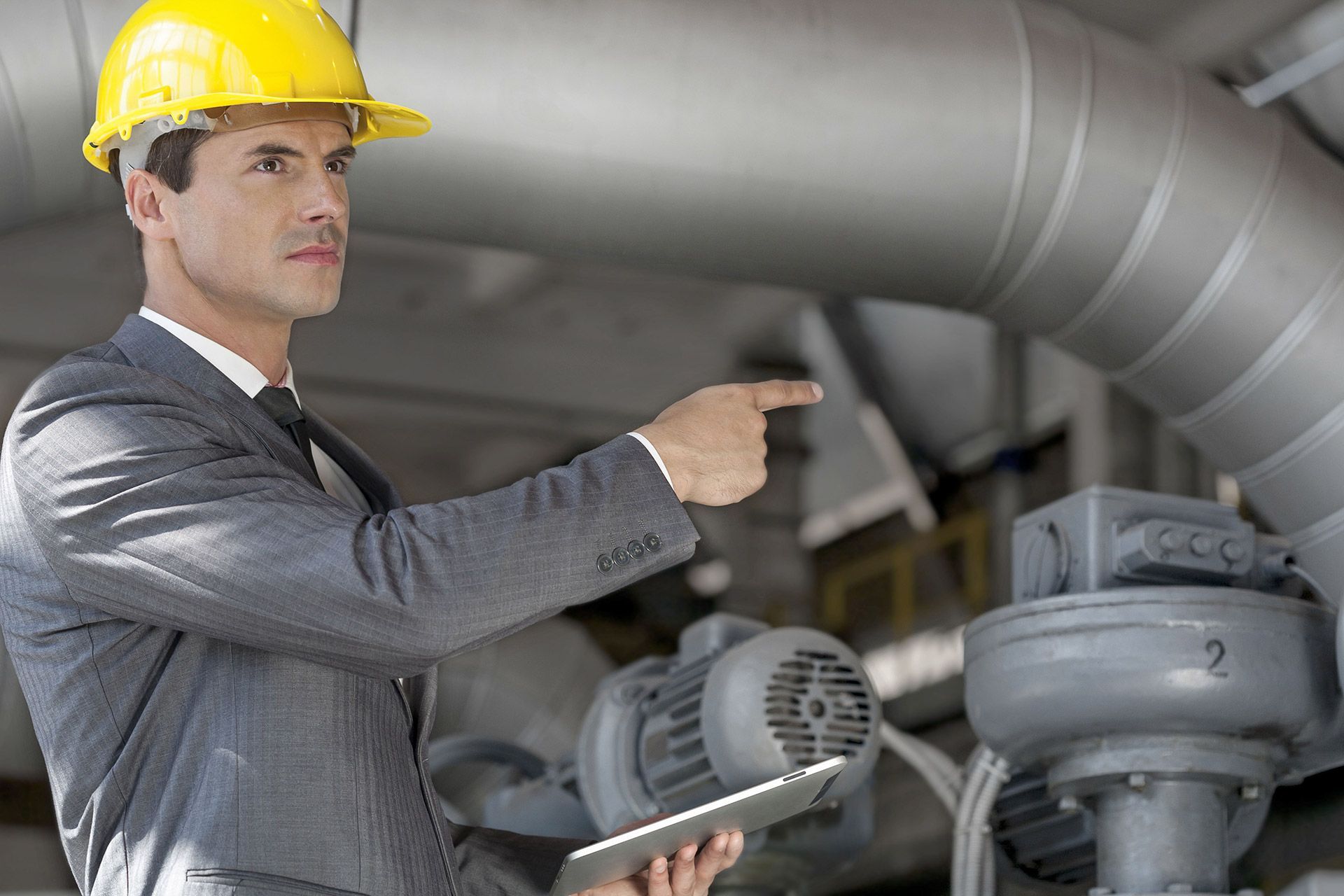 A man wearing a hard hat is pointing at something while holding a tablet.