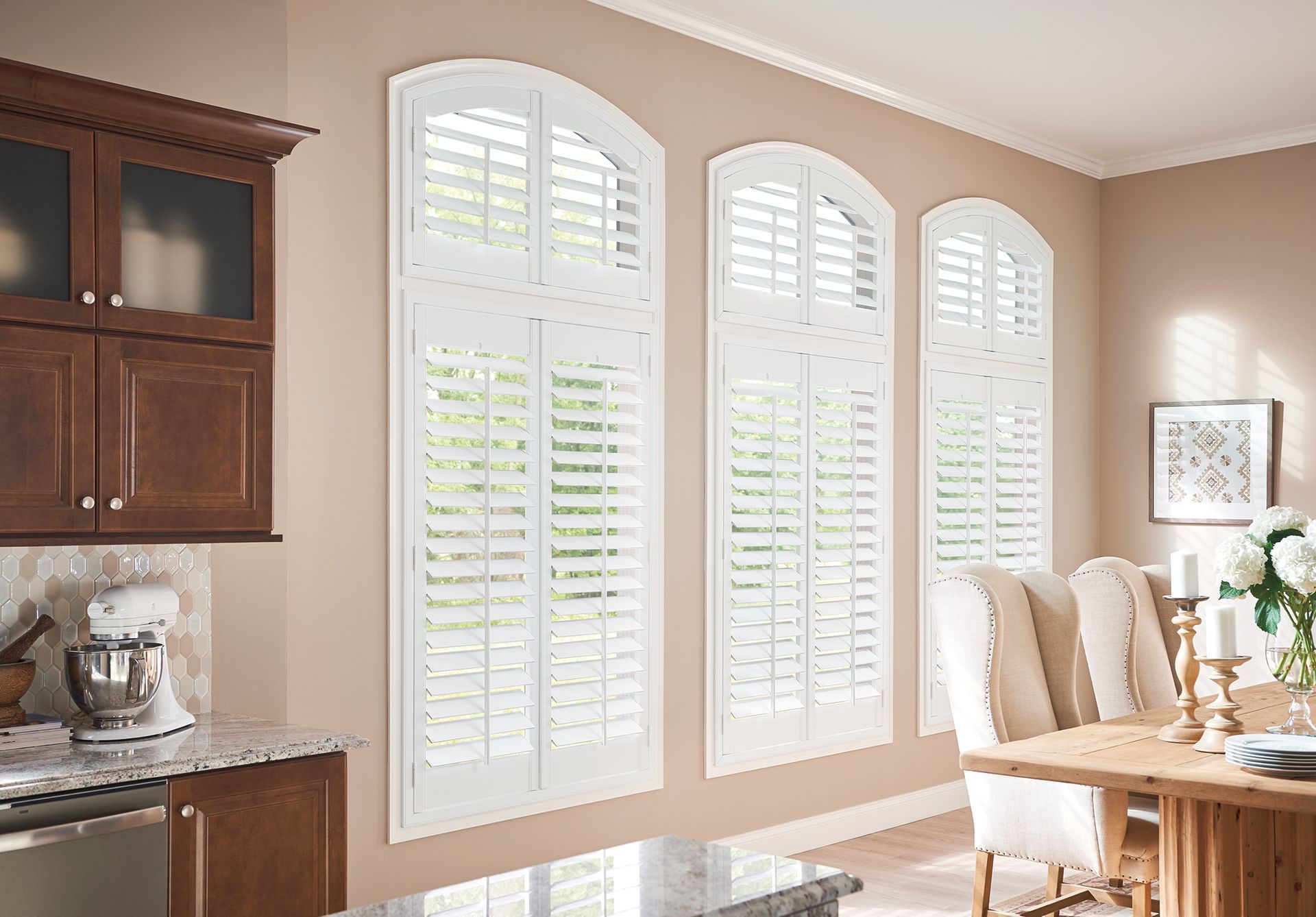 White arched shutters on tall windows in a dining room with tan walls and a wooden table.