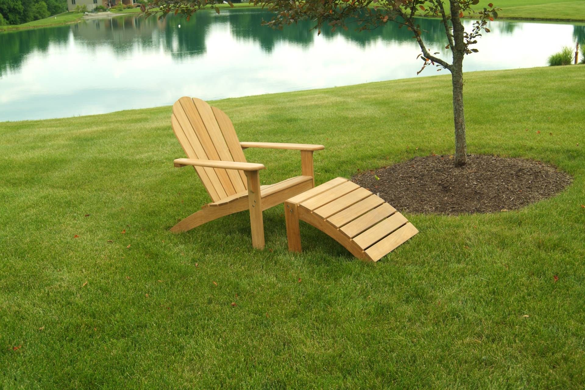 Wooden Adirondack chair and footrest on a grassy lawn next to a pond and small tree.