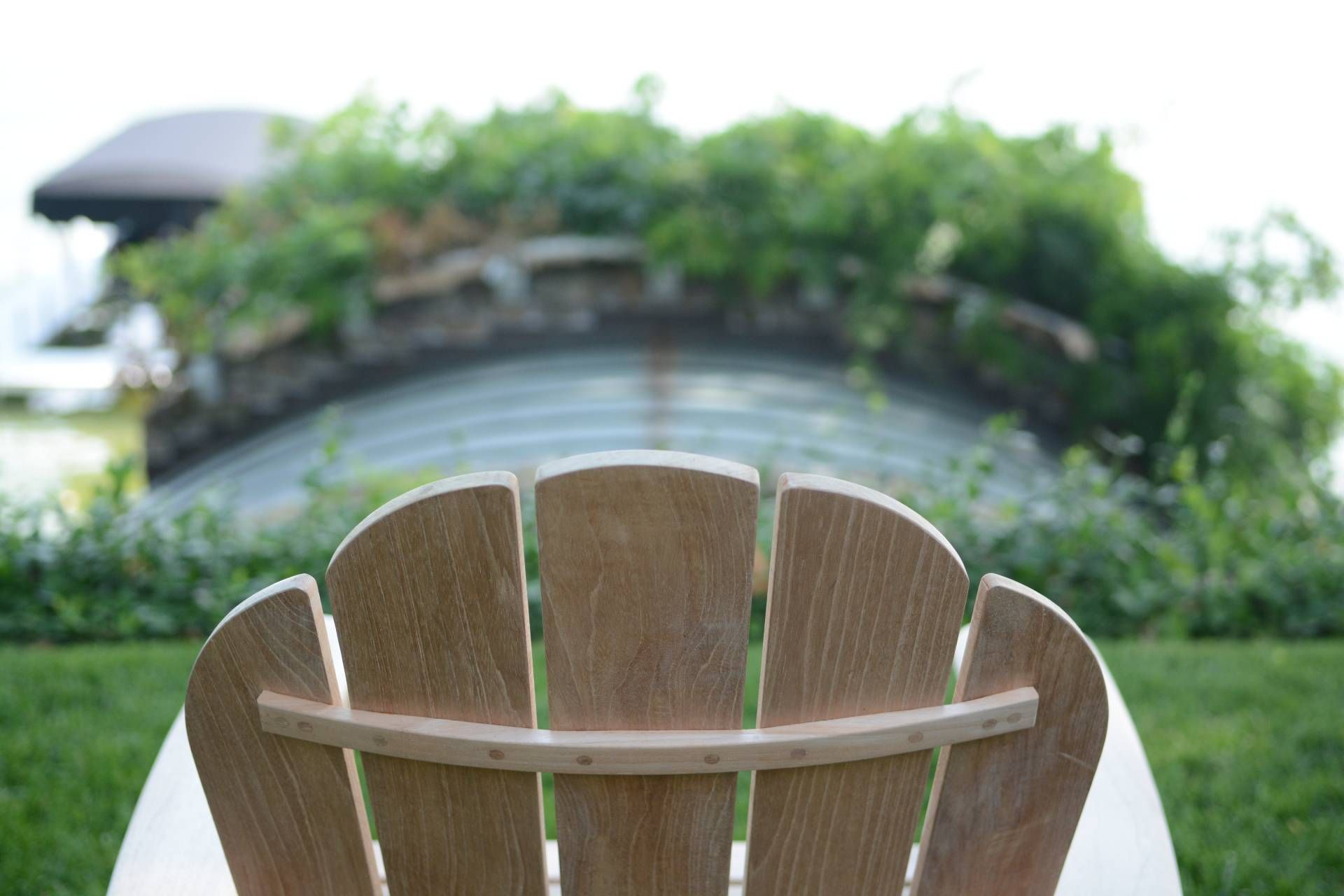Wooden Adirondack chair back with a curved backrest, overlooking a curved stone structure with greenery.