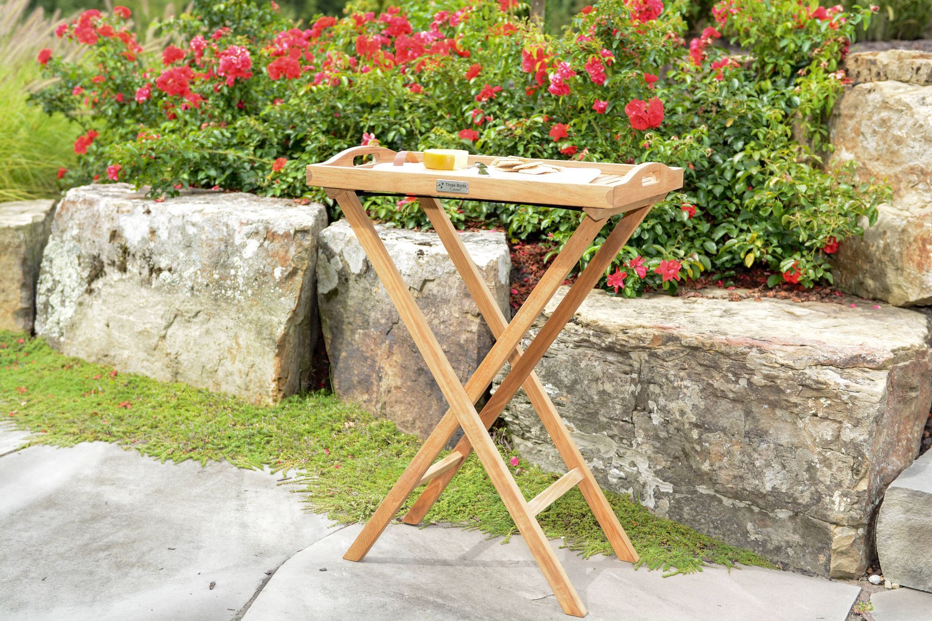 Wooden folding tray table outdoors, set in front of a stone wall with red flowers.