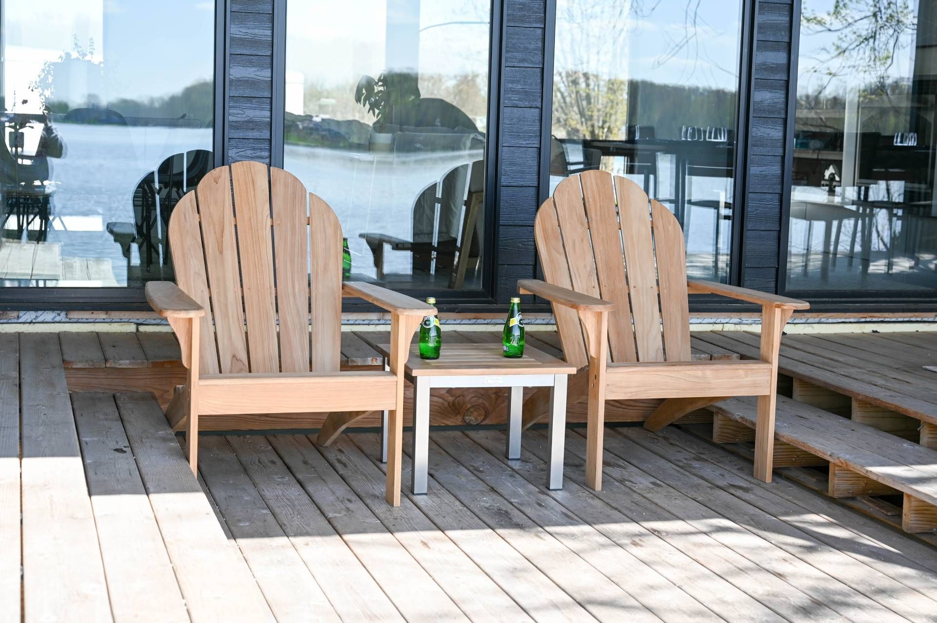 Two wooden Adirondack chairs and a small table with two bottles on a deck overlooking a lake.