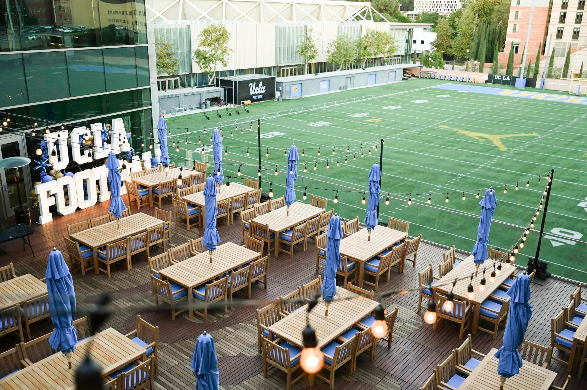 Outdoor dining area with tables, umbrellas, and a football field background. 