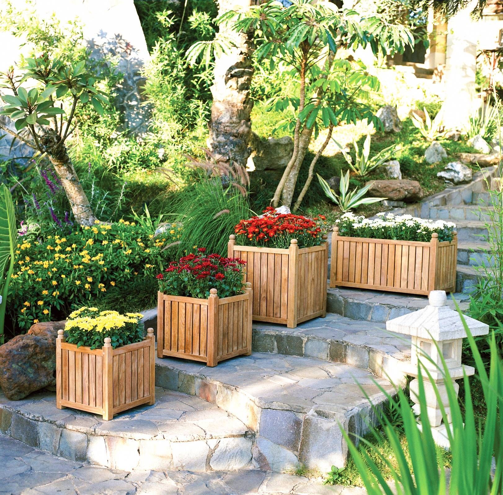 Four wooden planters filled with colorful flowers sit on stone steps in a garden.