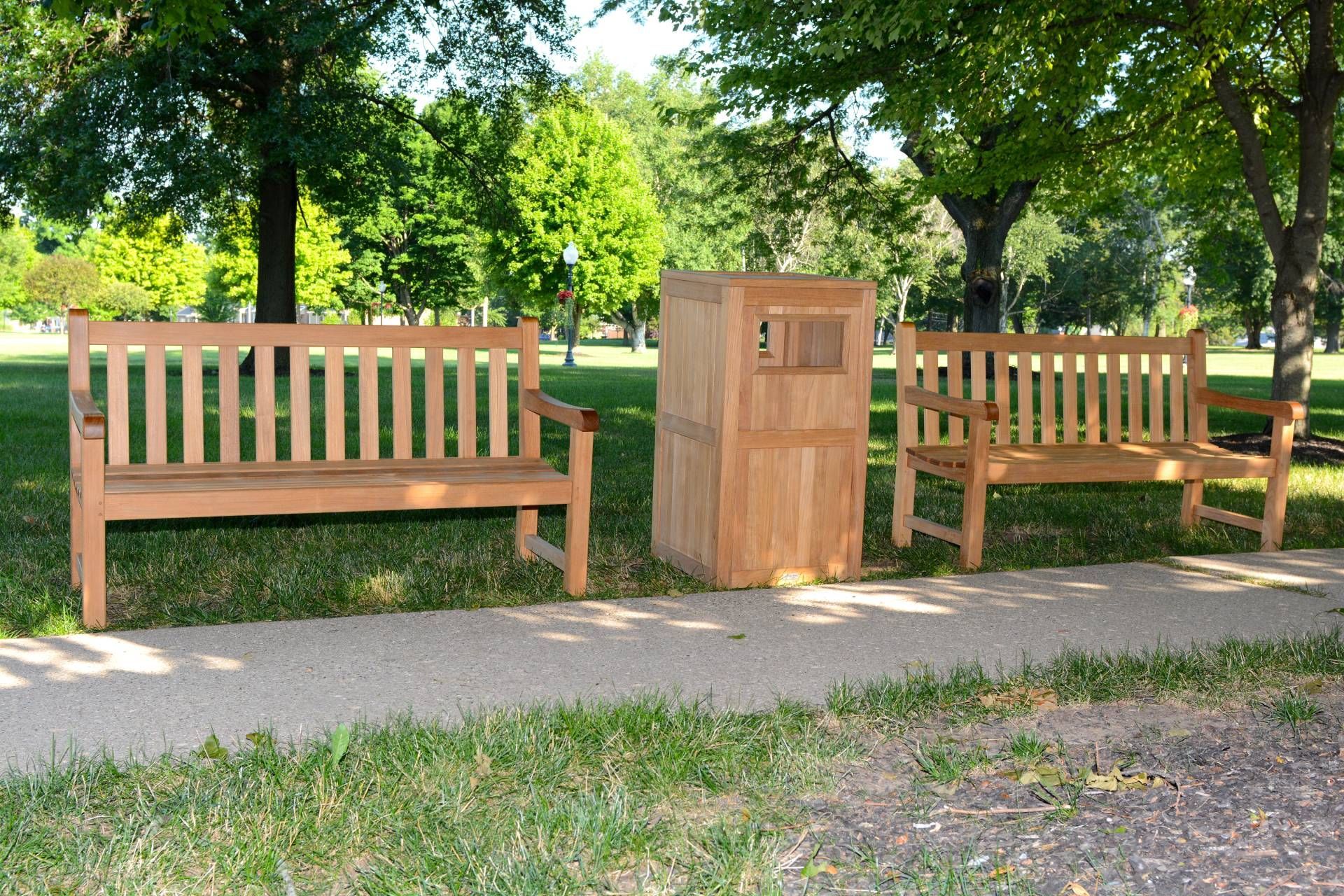 Two wooden park benches flanking a wooden box-like structure on a paved path in a grassy park. Trees in background.