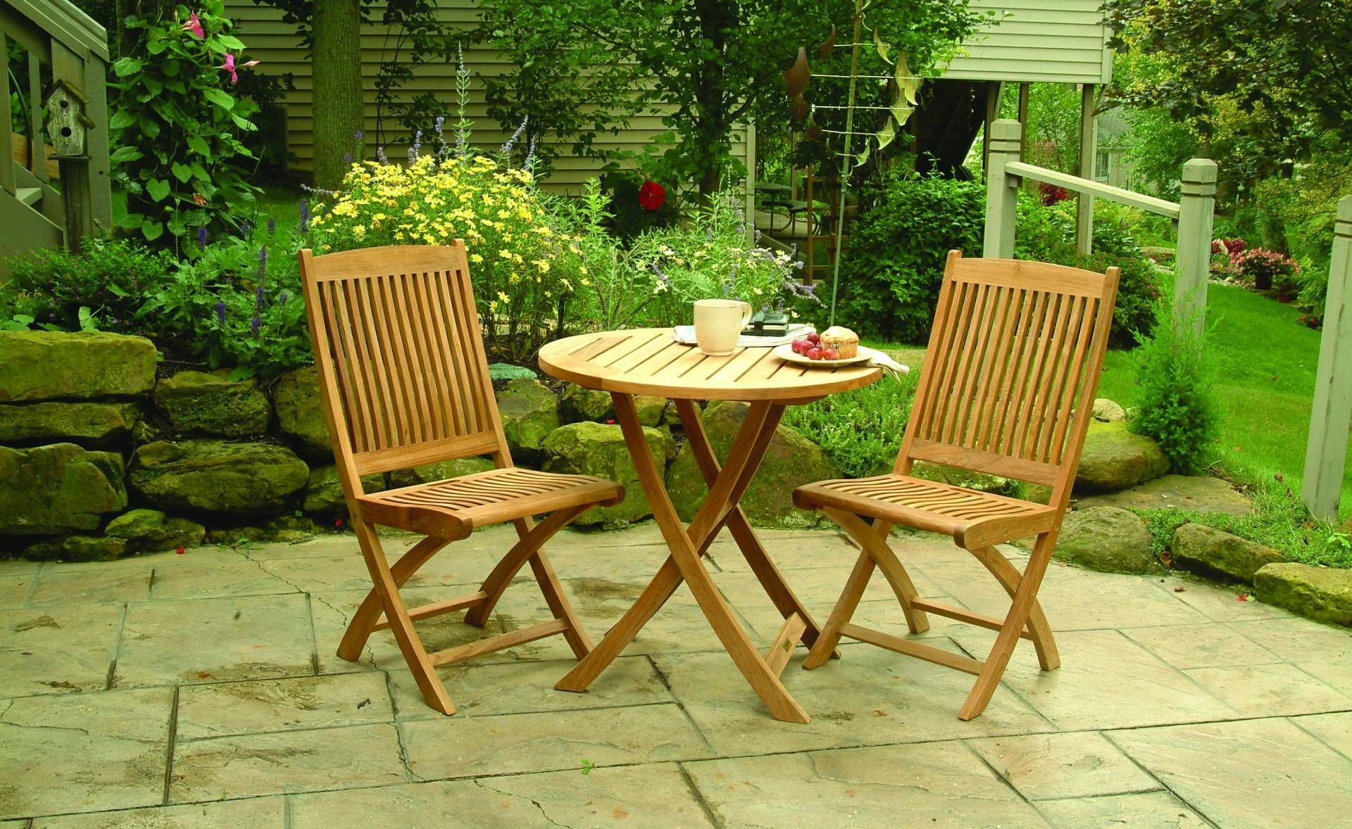 Two wooden chairs and a table on a patio, set for a meal. The scene is a garden with lush greenery.