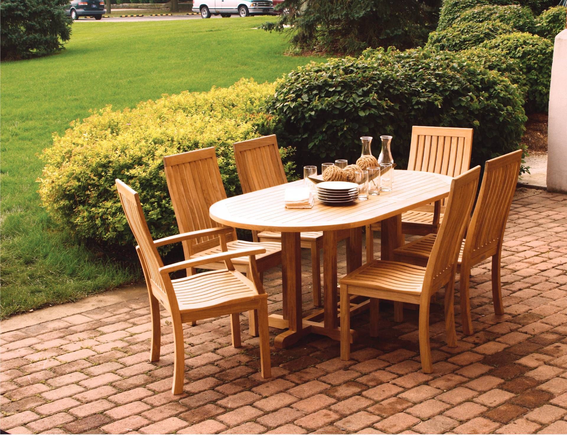 Wooden patio table with six chairs on a brick patio. Bottles and glasses sit on the table, with greenery in background.