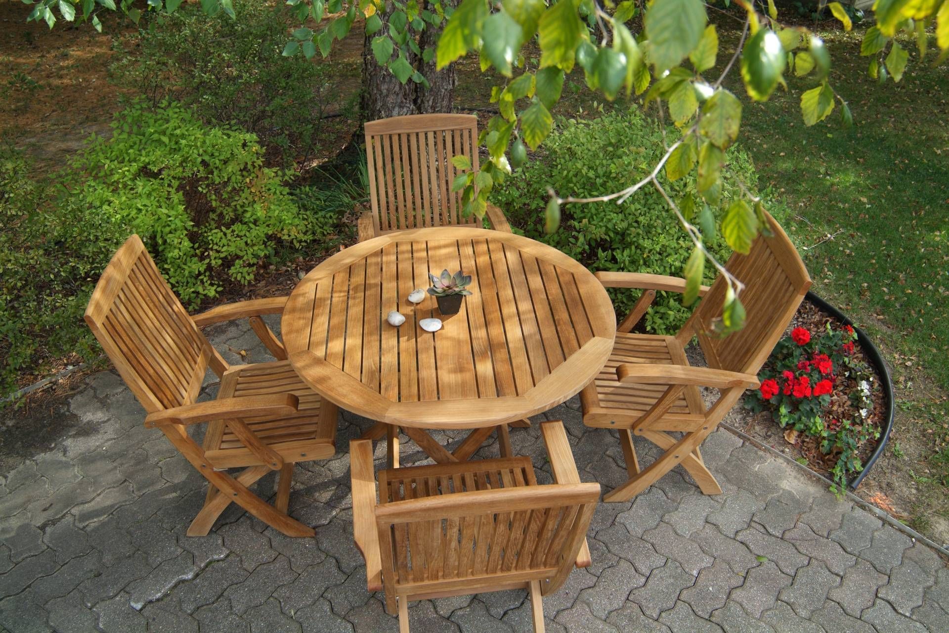 Wooden outdoor dining set beneath a tree, on a patio, surrounded by greenery.