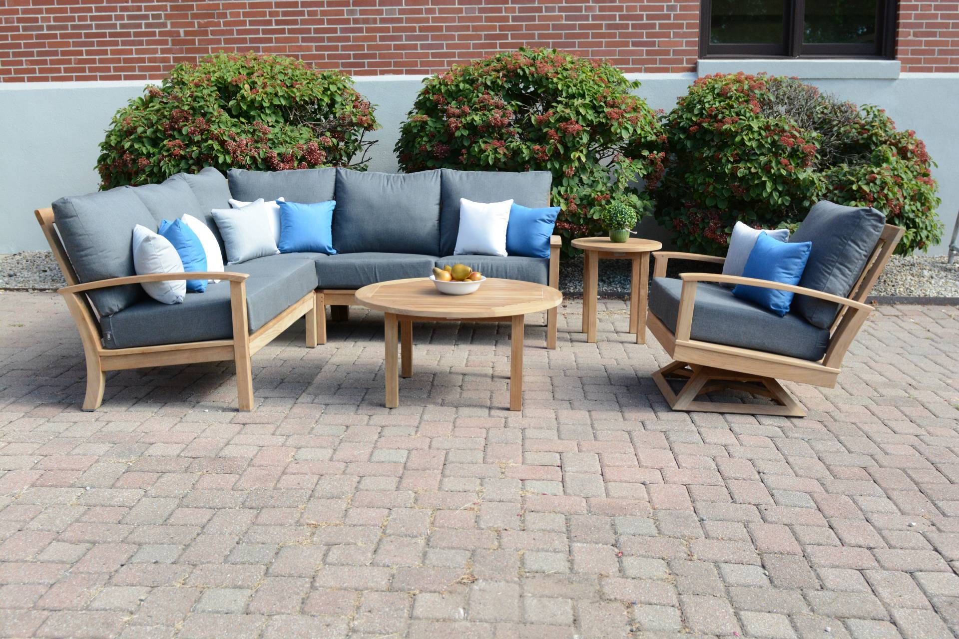 Outdoor teak furniture set with sectional, round coffee table, side tables, and swivel chair with blue cushions.