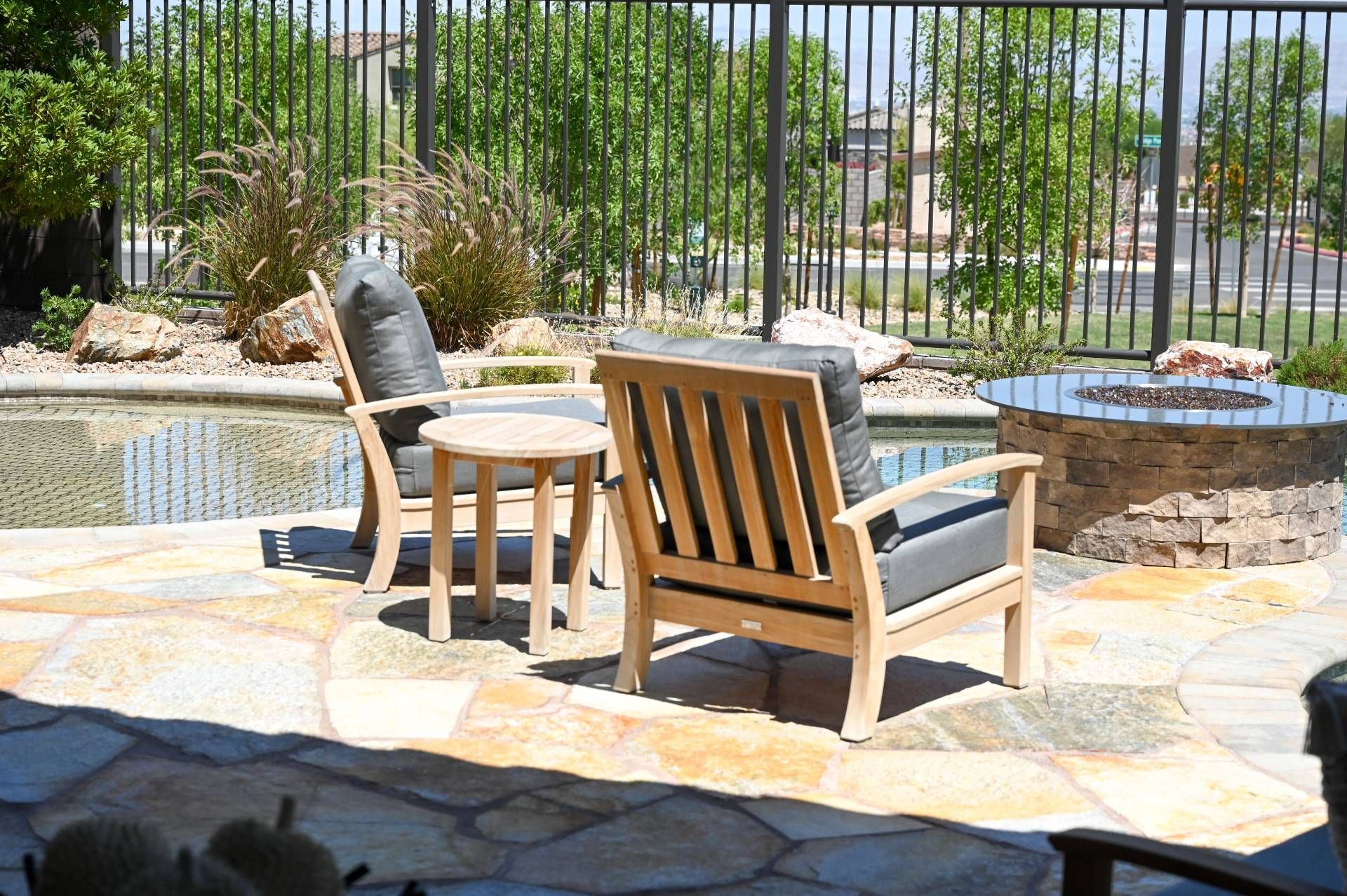 Two wooden chairs with cushions and a small table on a stone patio near a fire pit, with a fence and greenery in the background.
