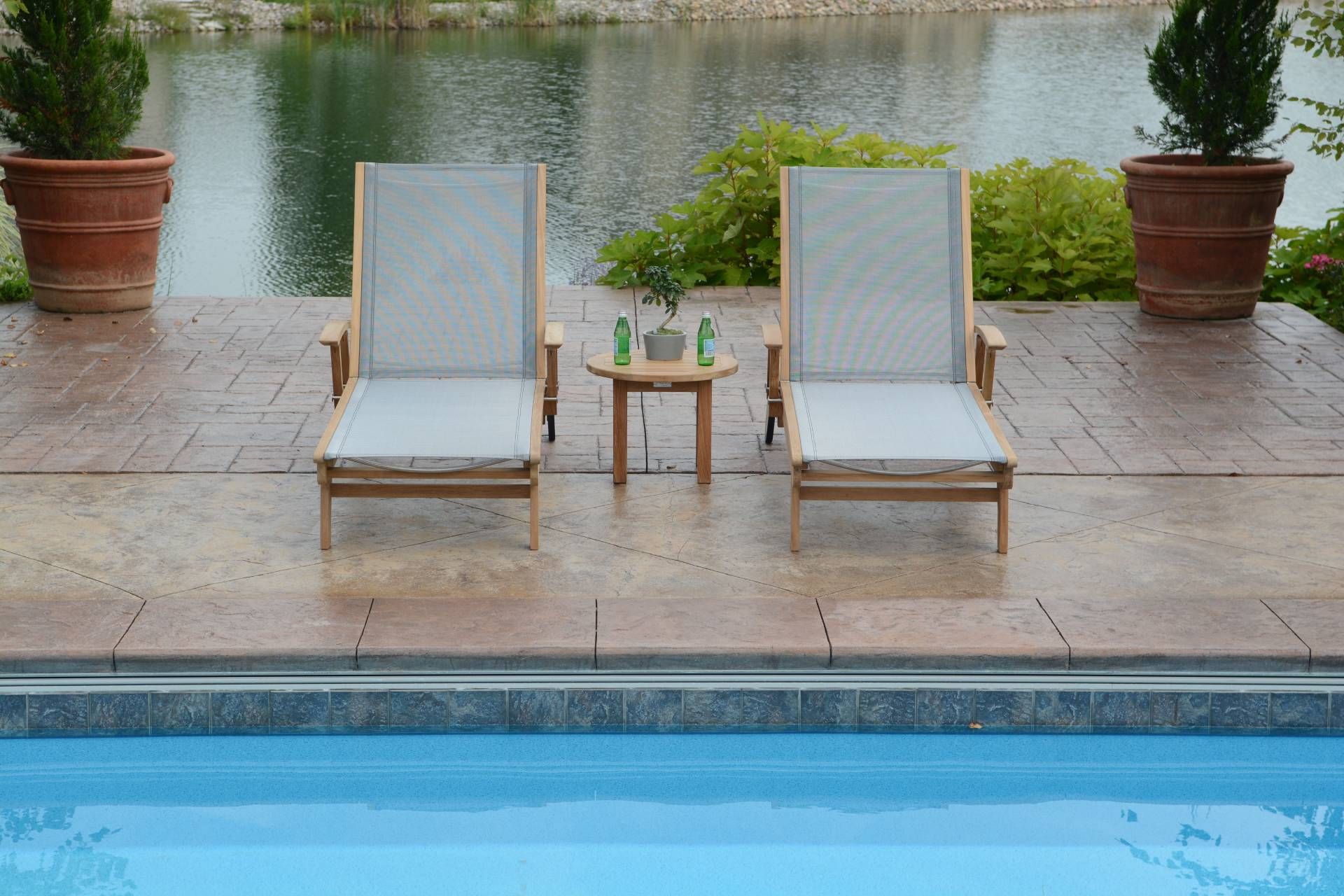 Two wooden lounge chairs and small table beside a pool, overlooking a lake.