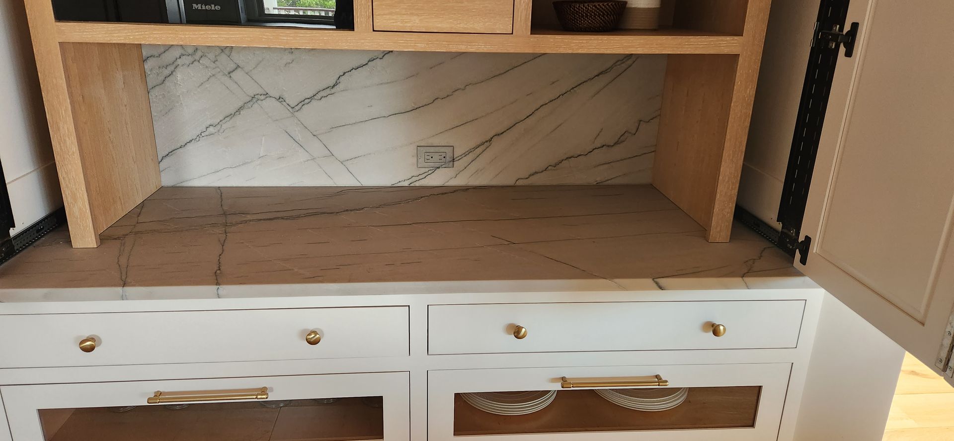 White cabinet with wooden interior and marble backsplash, gold knobs, and open drawers.