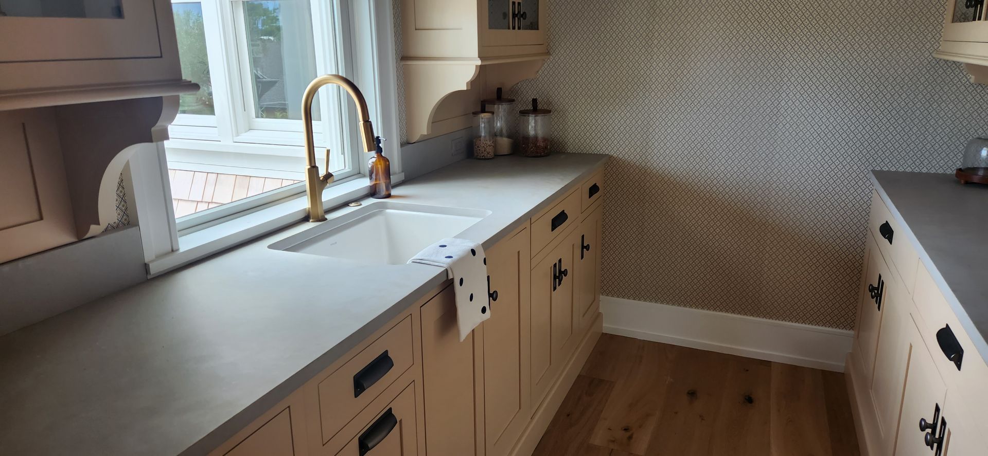 Kitchen with light-colored cabinets, countertop, and gold faucet; wood flooring and patterned wallpaper.