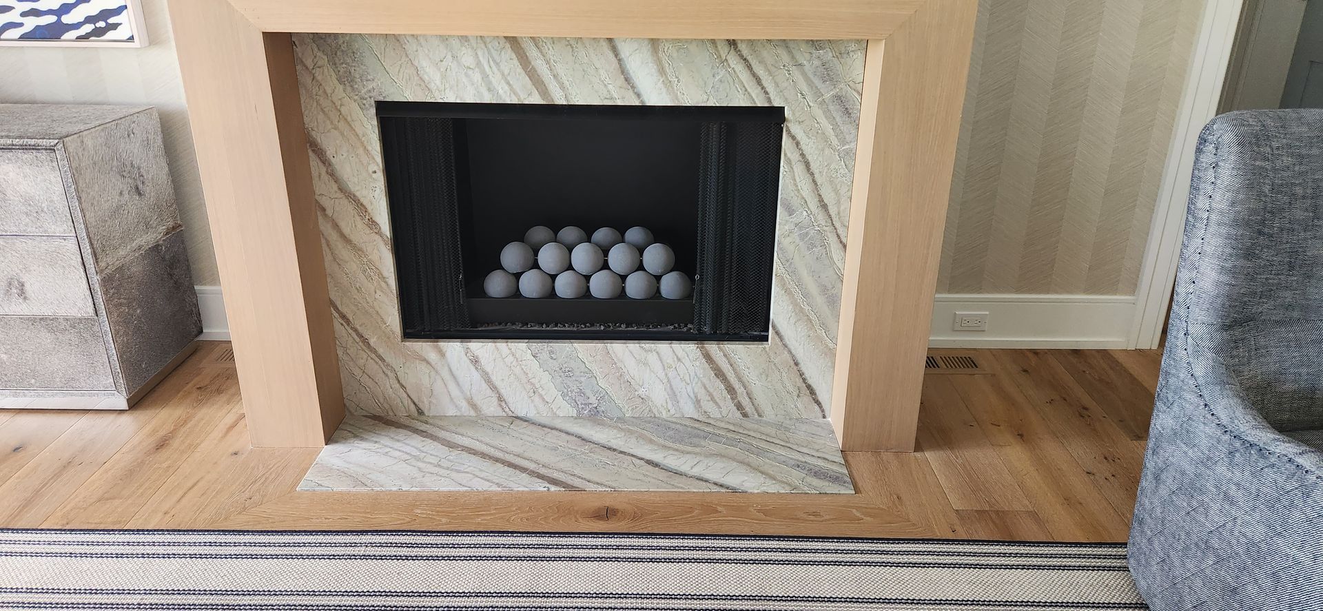 Fireplace with gray stone surround and light wood mantel, on wood floor, gray spheres in firebox.