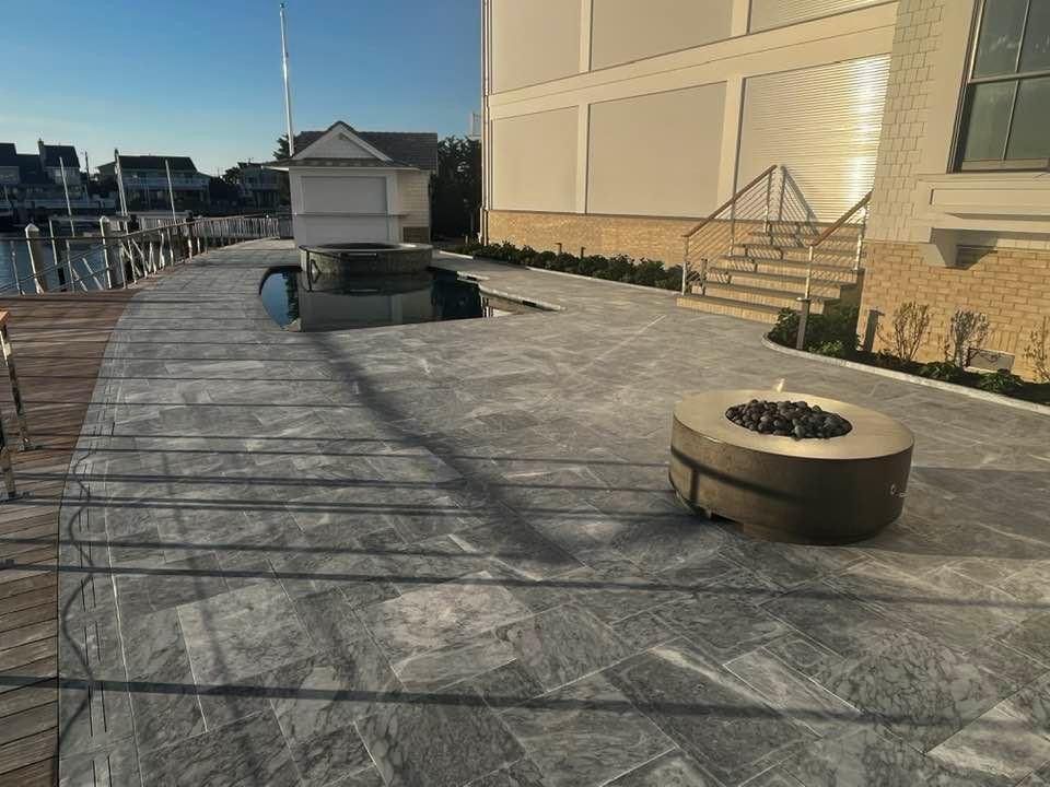 A concrete patio by water's edge with a fire pit, stairs, and building. Blue sky.