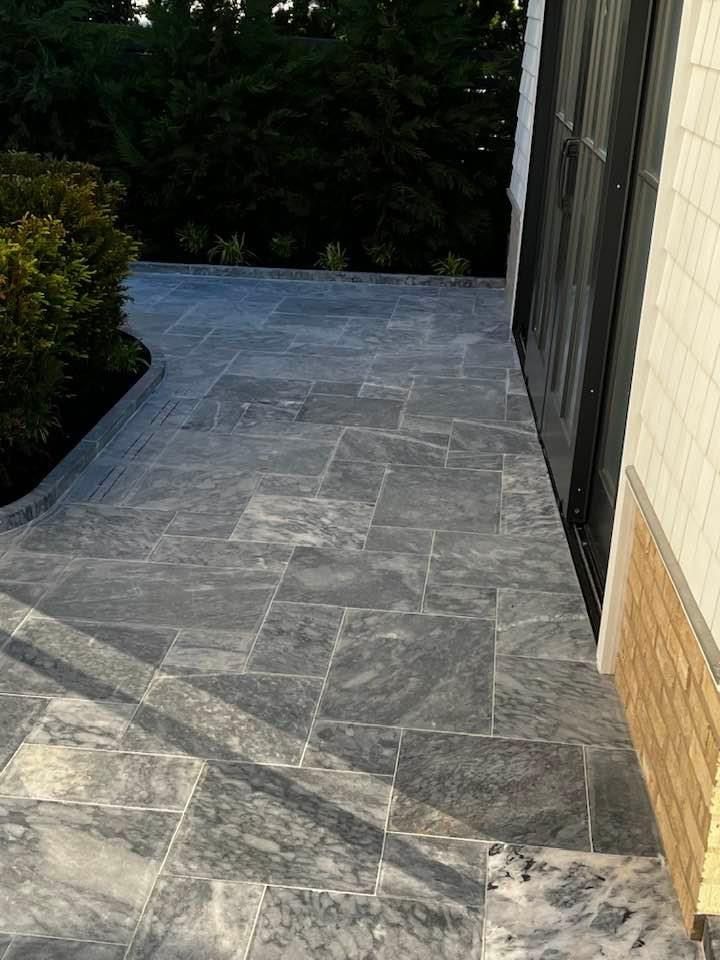 Grey stone patio with irregular rectangular tiles next to a building and green hedge.