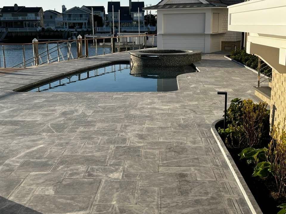 Patio with a pool and hot tub overlooking a waterway, grey paving, sunny day.